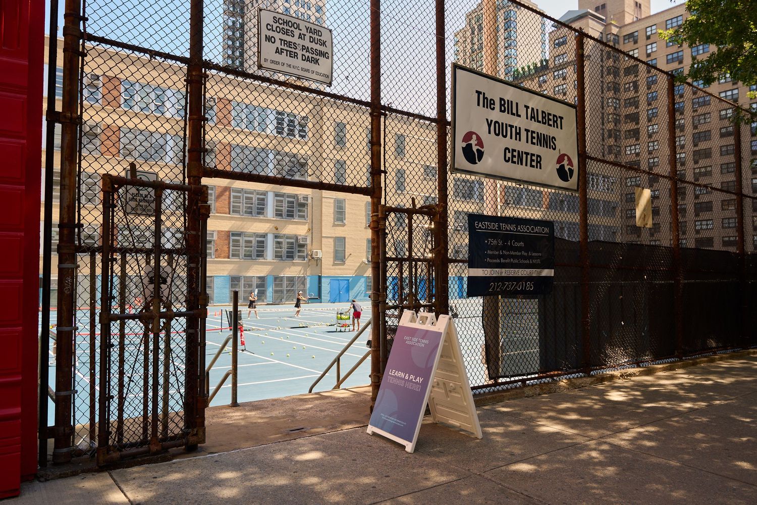Fenced tennis court surrounded by tall apartment buildings in an urban setting.
