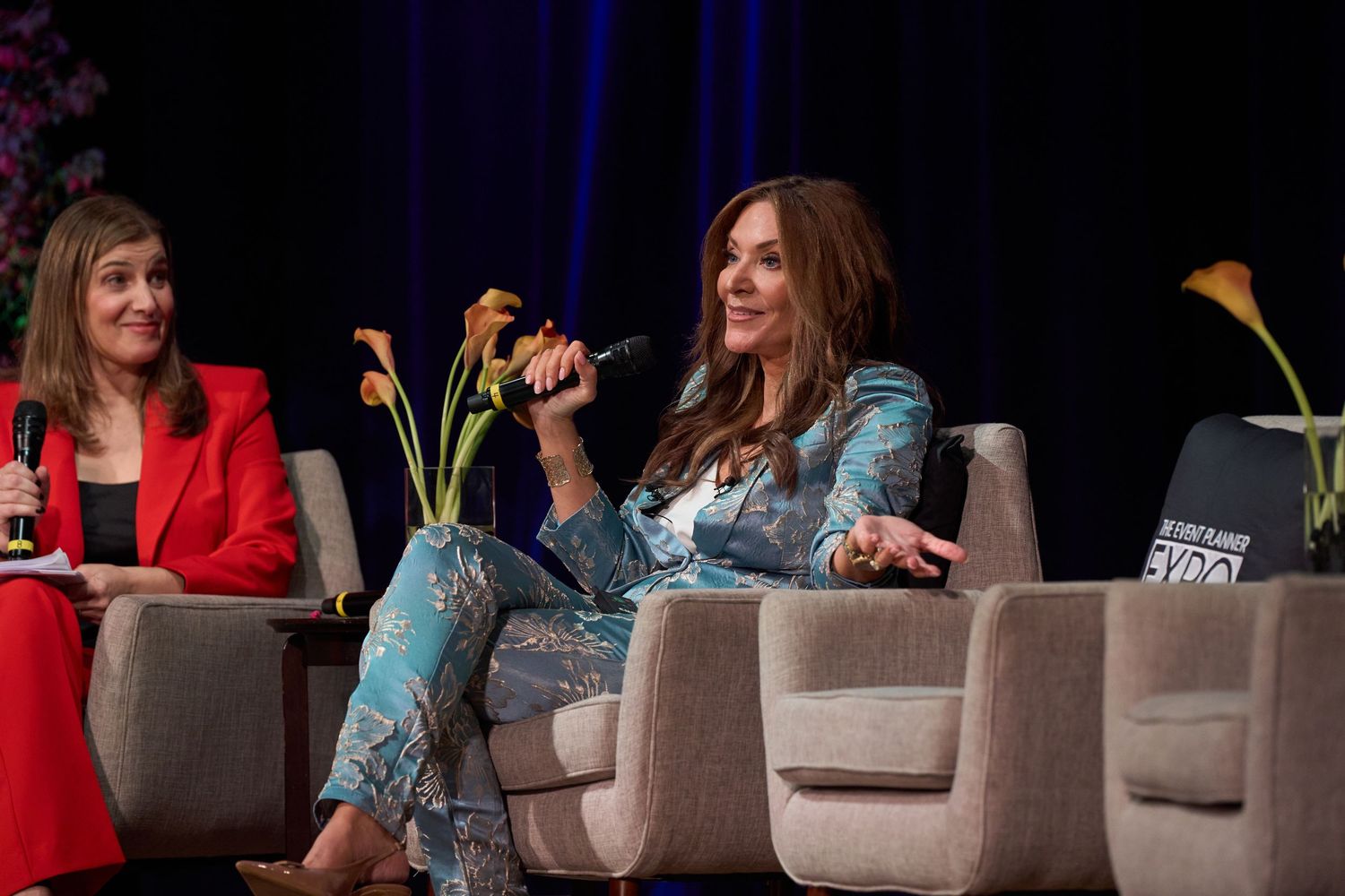 Speakers seated in gray armchairs on stage during a panel discussion event with blue curtain backdrop.