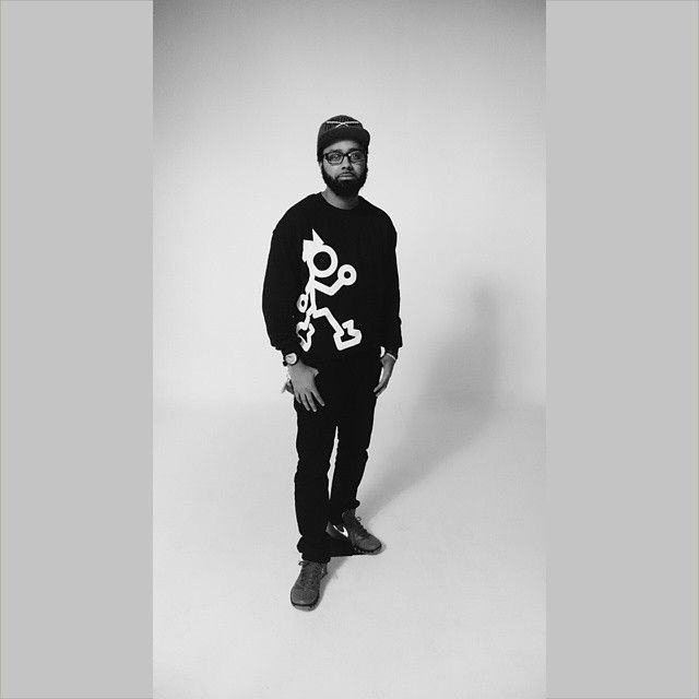 Black and white portrait of person wearing dark sweater with graphic design standing against plain background.