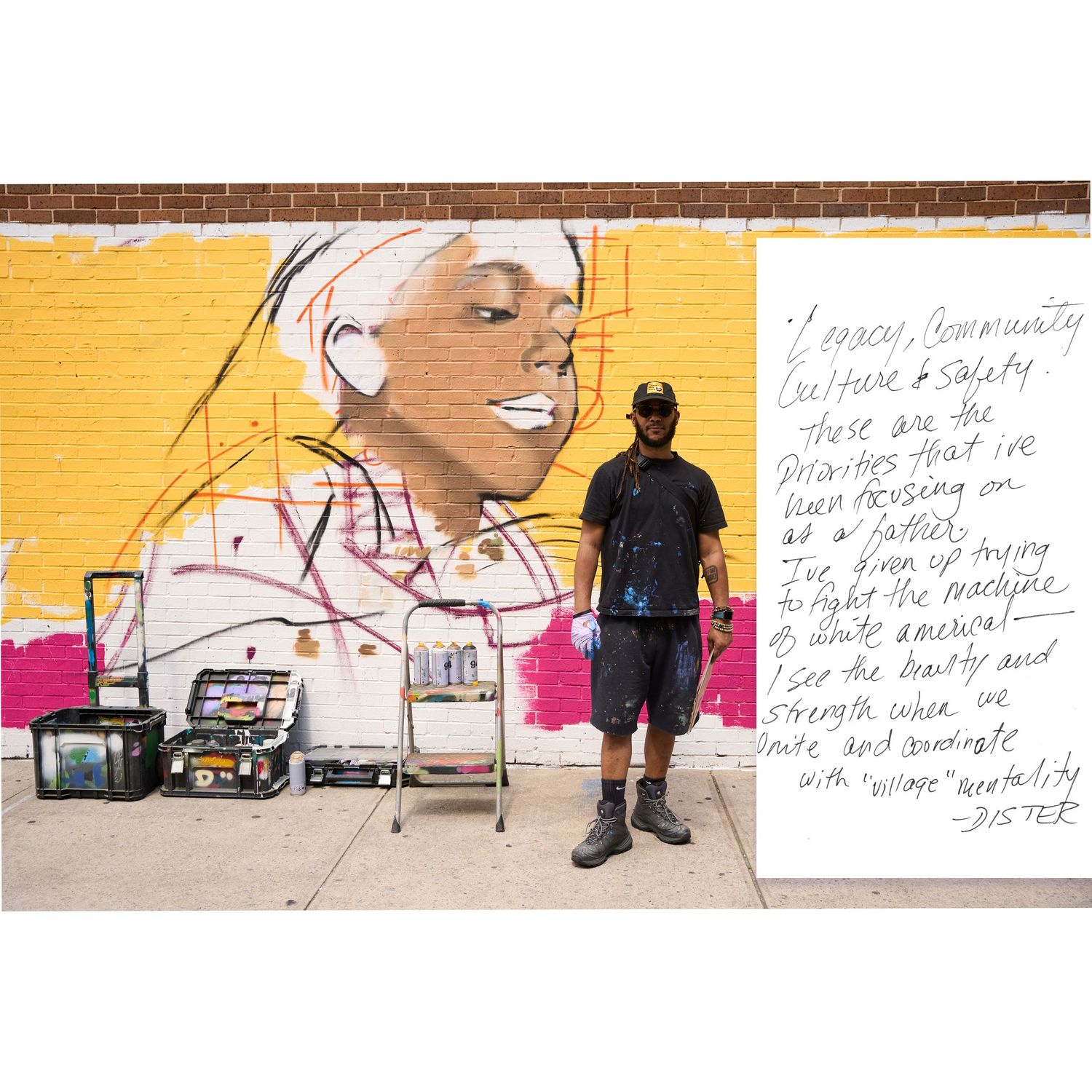 Artist in black clothing stands in front of a colorful mural-in-progress on a yellow wall with painting supplies nearby.