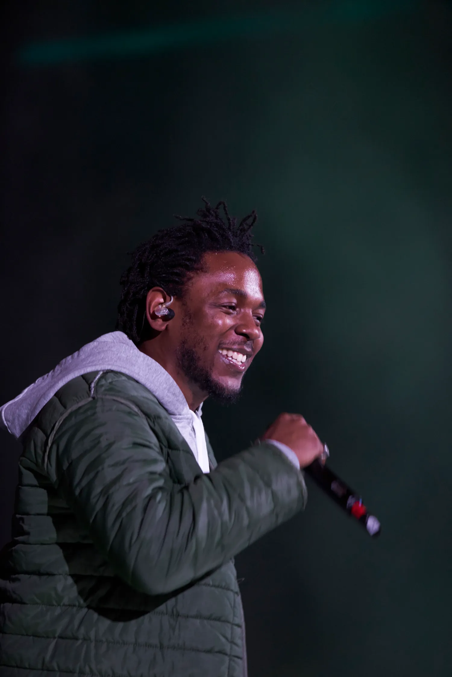 Performer singing with enthusiasm in dark olive quilted jacket against a dramatic teal-tinted stage backdrop.