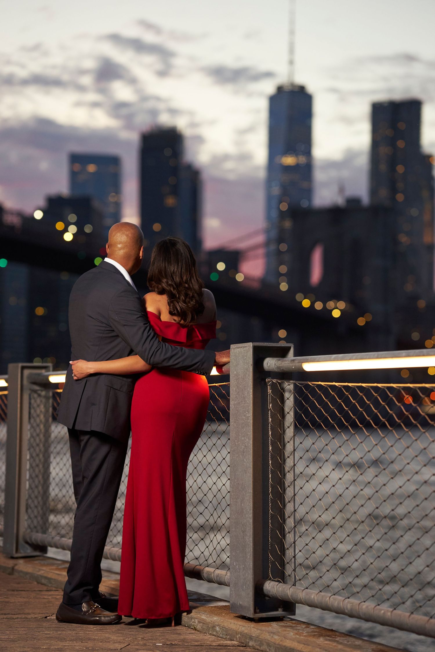 A couple embraces while looking at a city skyline at sunset from an observation deck with metal railings.