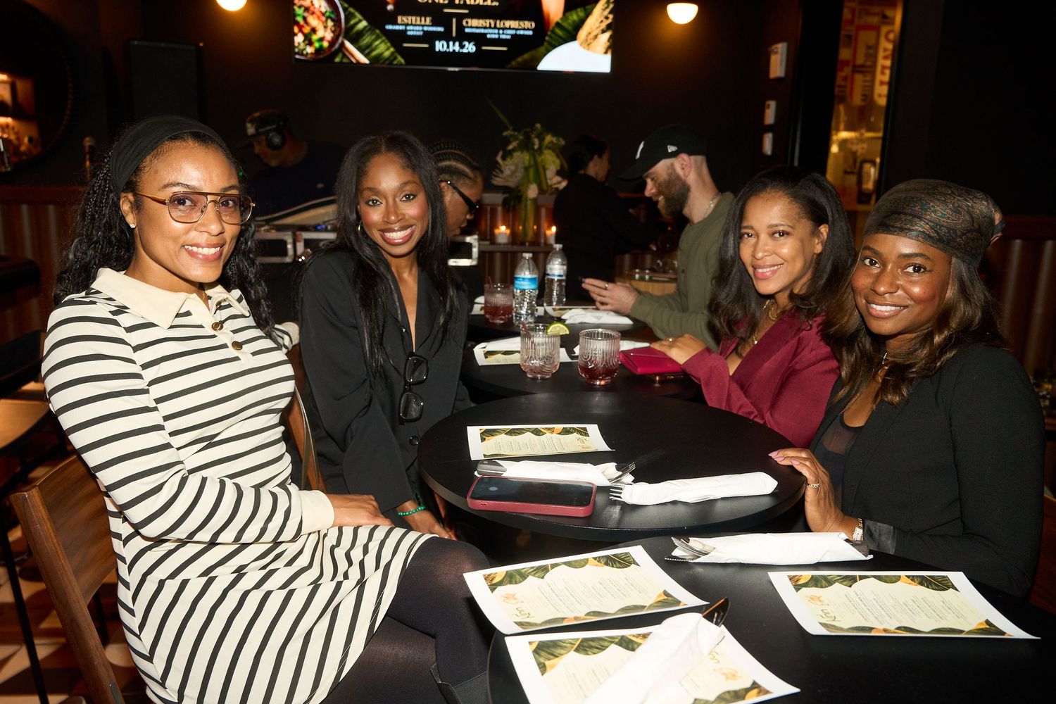 Four smiling women in stylish outfits pose together at a restaurant table set with menus, drinks and plates in a warmly lit dining room.