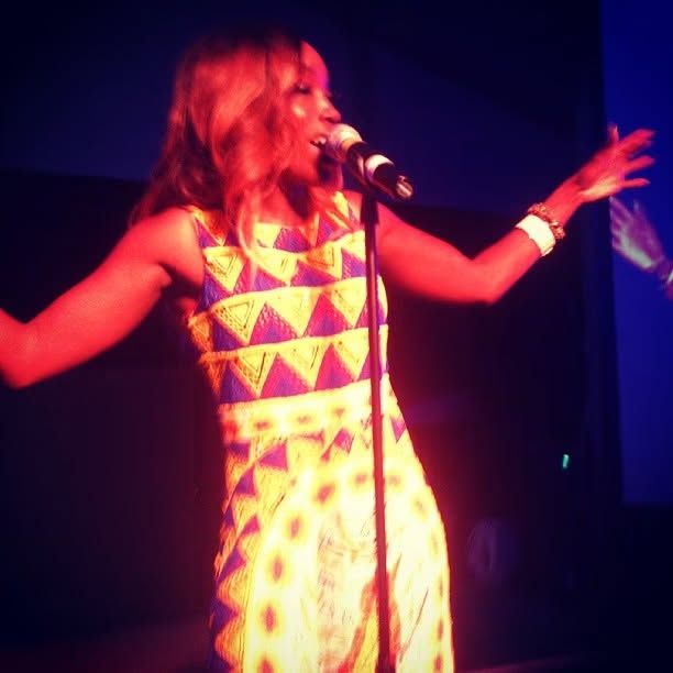 Female singer in a colorful geometric-patterned dress performs on stage with arms outstretched while singing into a microphone.
