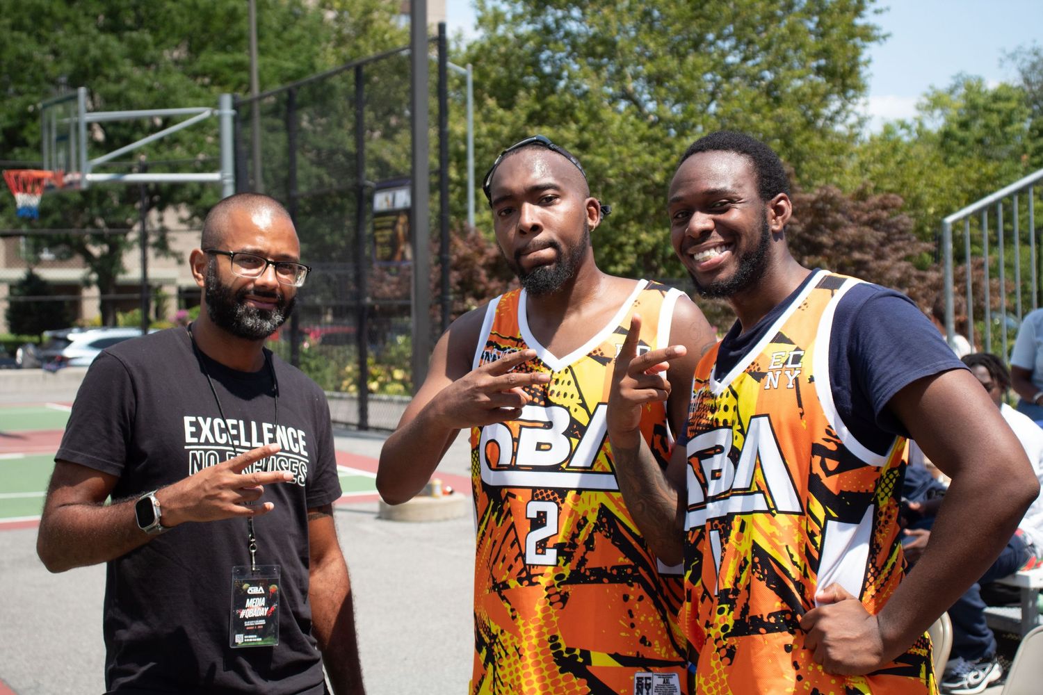 Three athletes in basketball jerseys stand together smiling on an outdoor court on a sunny day.