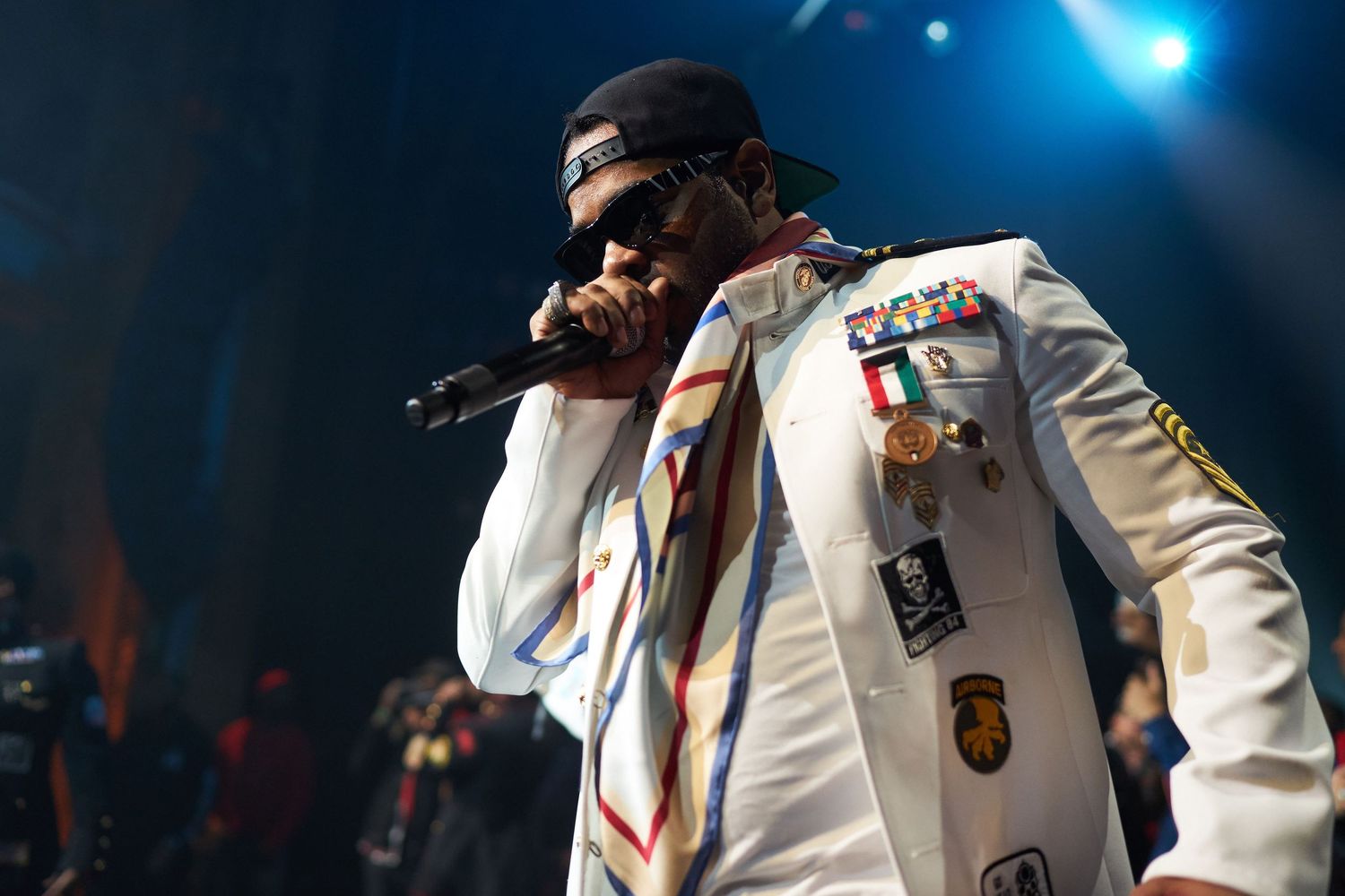 Performer in white embellished jacket sings into microphone on stage with blue lighting in background.