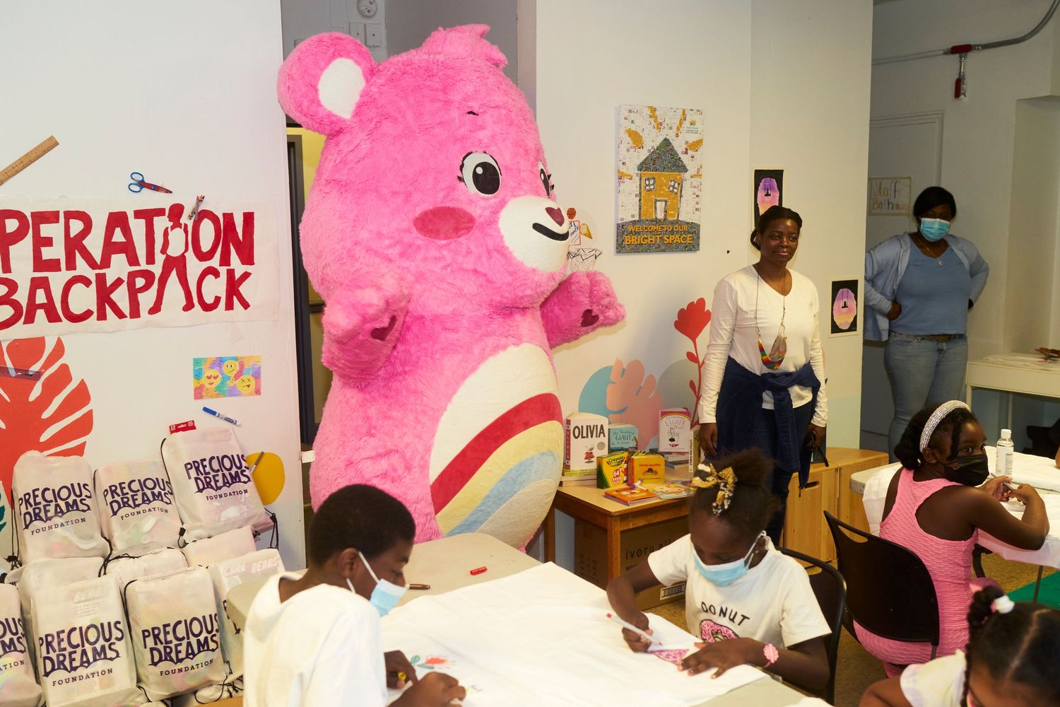 A bright pink Care Bear plush mascot poses at a promotional event with posters and signs in the background.