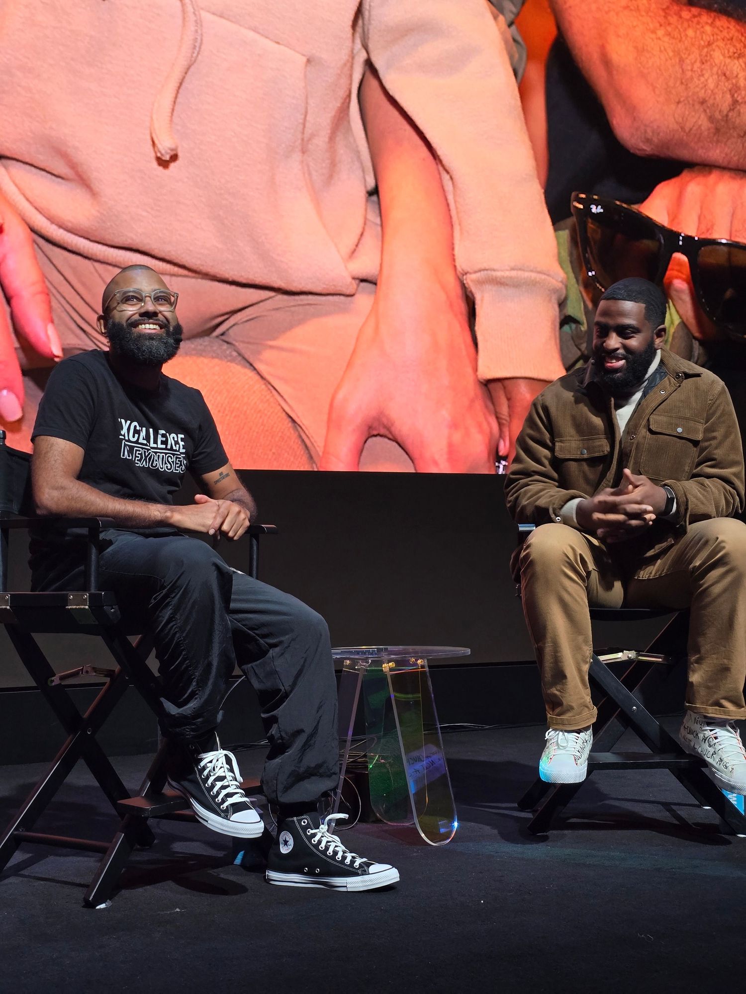 Casual footwear detail showing several pairs of legs seated in chairs, with one person wearing black sneakers against an orange background.