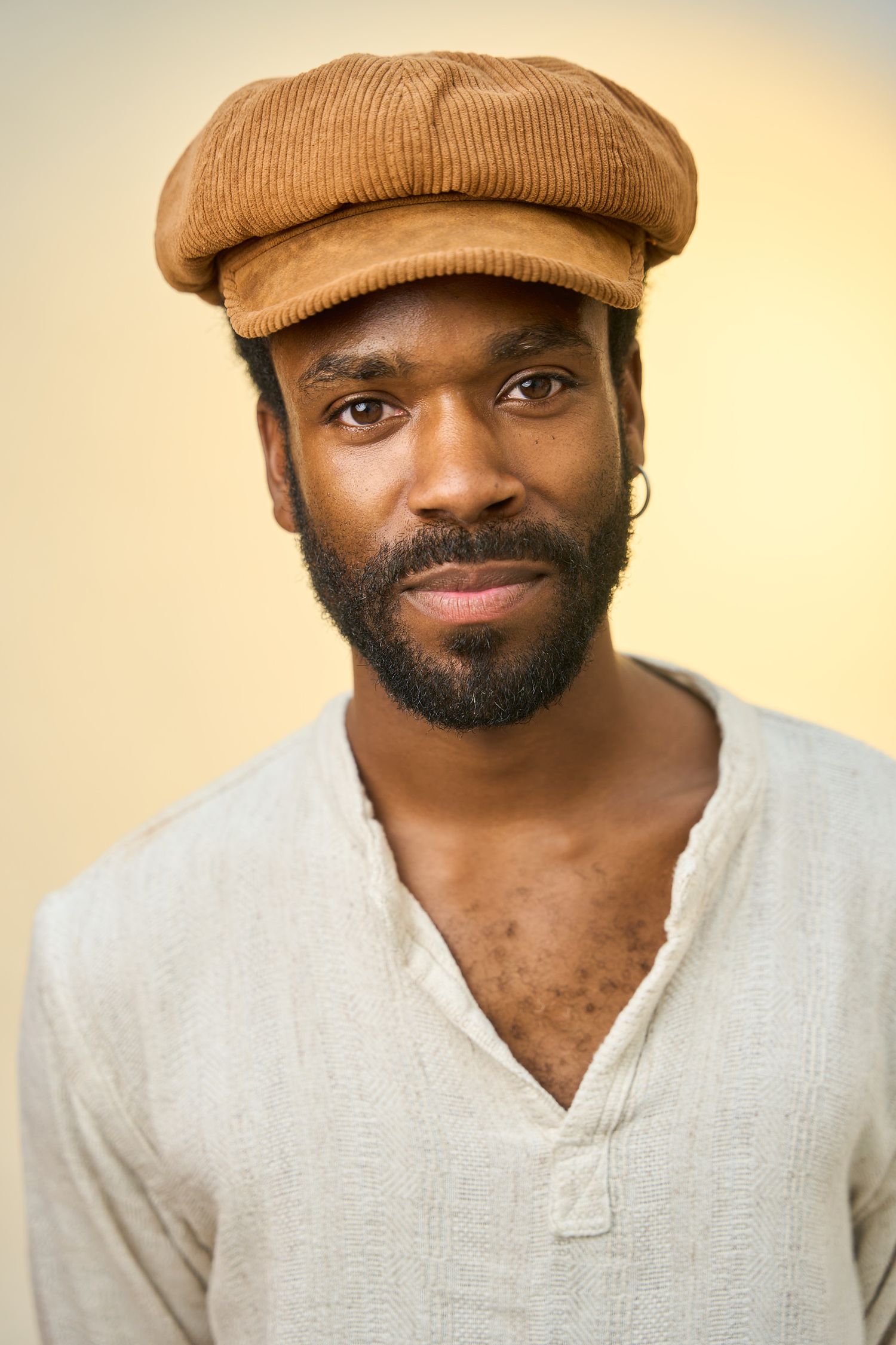 Portrait of a bearded Black man wearing a brown corduroy flat cap and linen shirt, looking at camera.