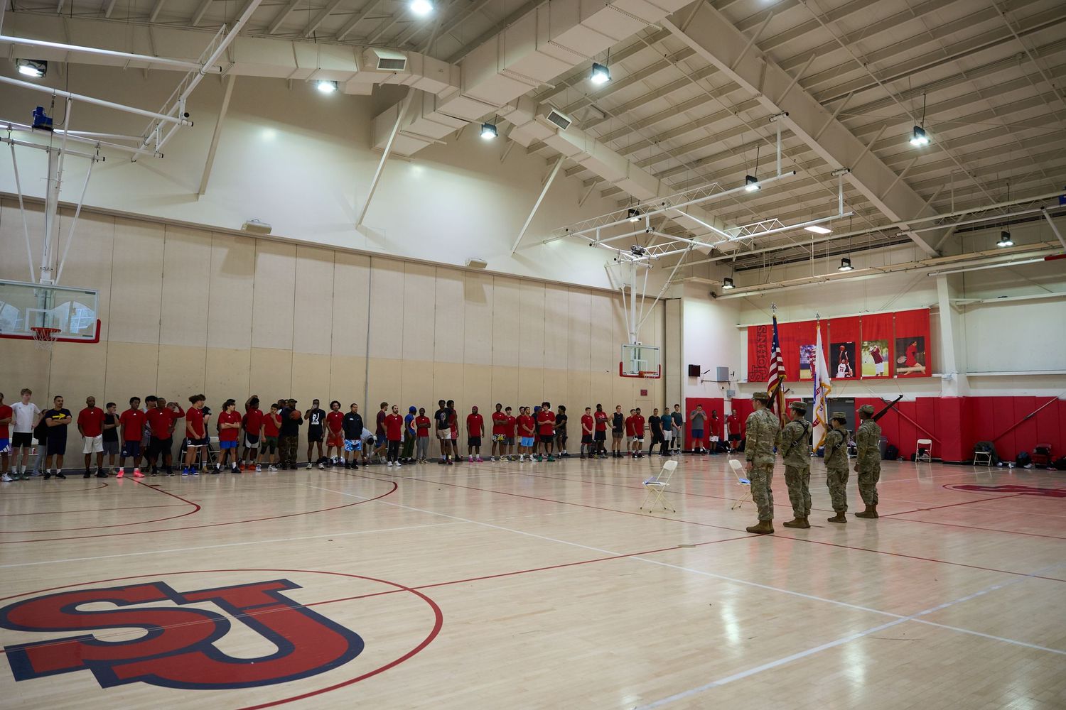 Wide angle view of a spacious indoor basketball gymnasium with red walls and bright overhead lighting.