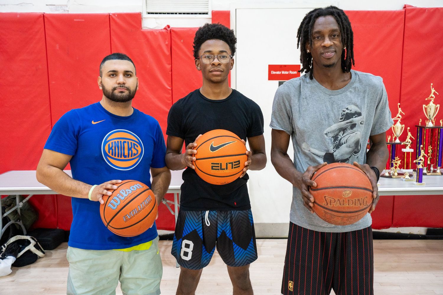 Basketball players line up in a gym with red walls and hold basketballs while wearing athletic attire.