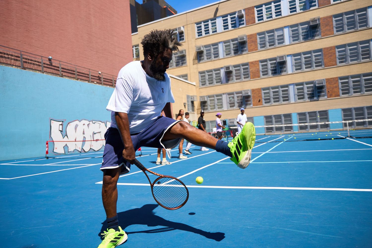 Players practicing tennis drills on a blue court with a brick building in the background.