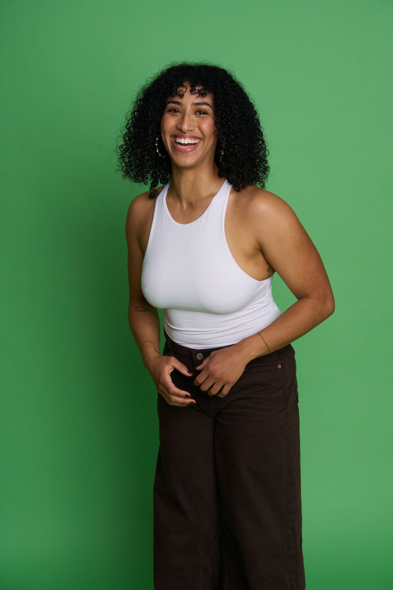 Smiling woman with curly hair wearing a white tank top and brown pants poses against a green background.