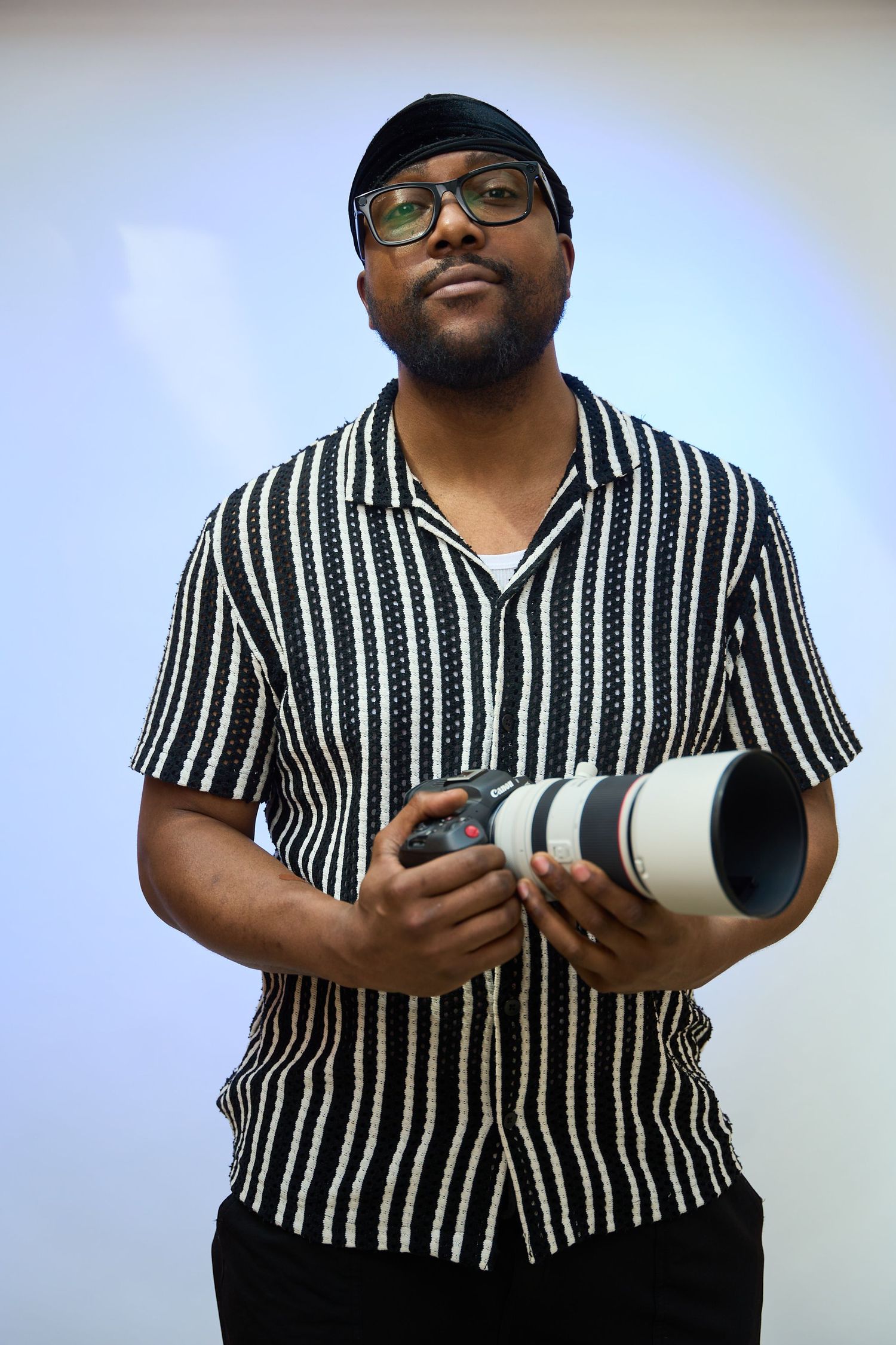 A man with glasses and a black cap holds a professional camera with a telephoto lens, wearing a striped shirt.