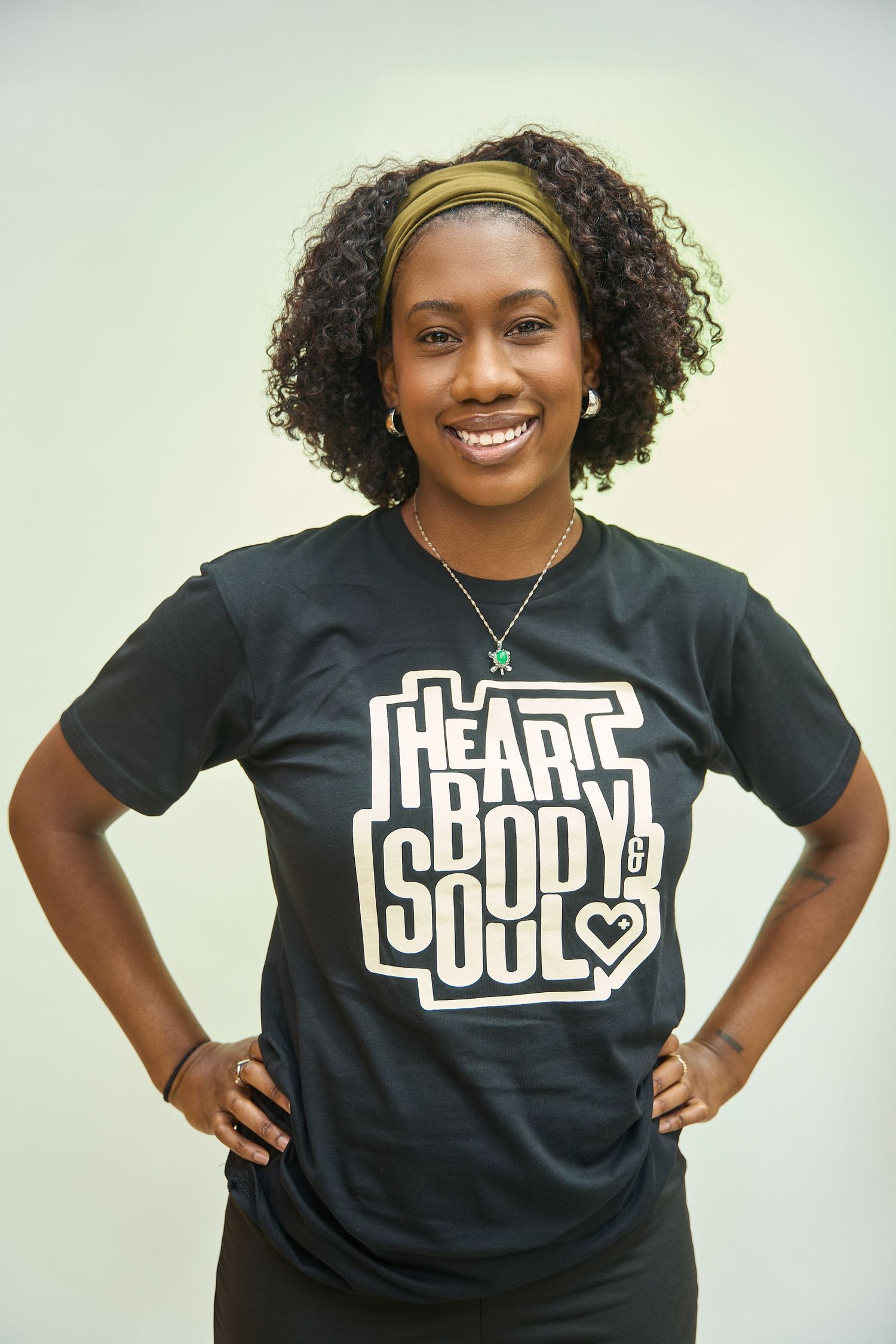 Smiling woman wearing a black Heart Body & Soul graphic t-shirt with hands on hips against a light background.