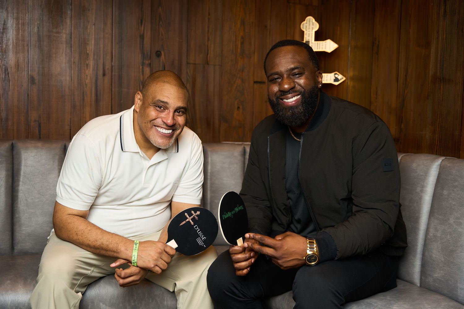 Two men in casual attire sit on a gray couch sharing laughs while having a conversation in a wood-paneled room.