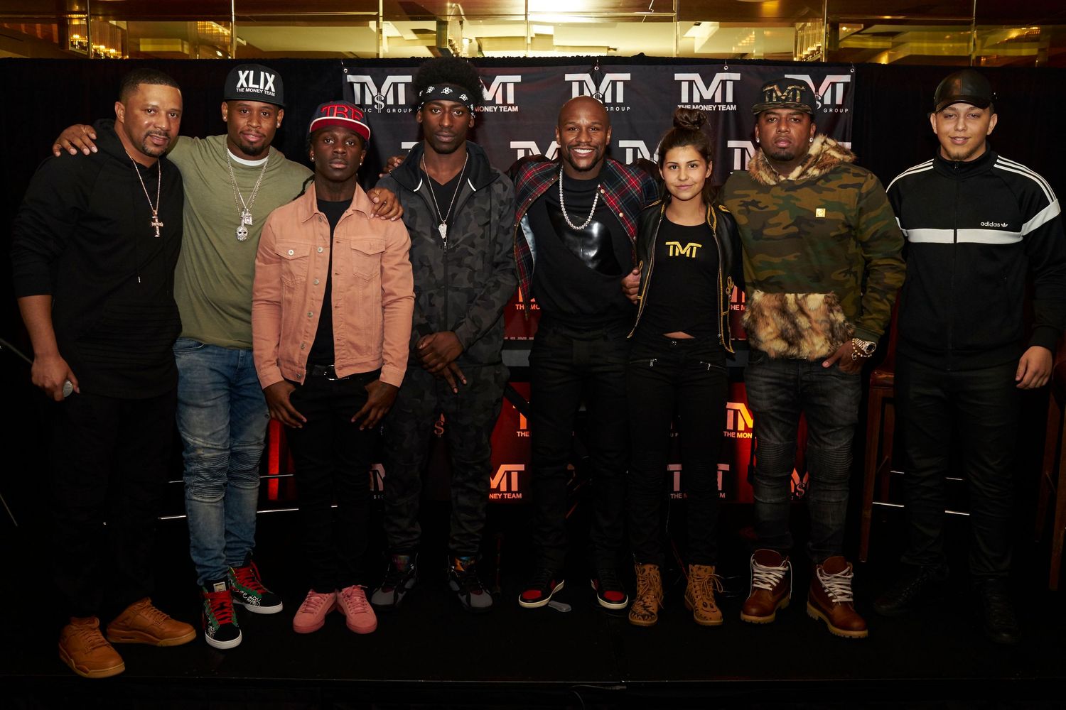A group of people pose together in casual attire in front of a dark backdrop with TMT branding visible.