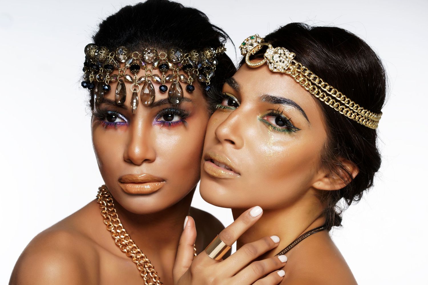 Two models wearing gold jewelry and dramatic makeup with metallic eyeshadow and glowing skin pose together.