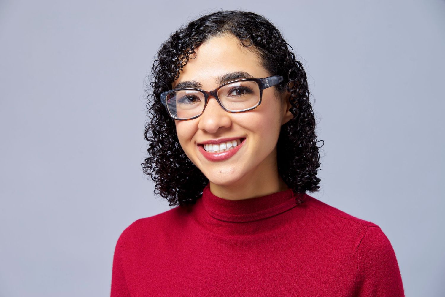 A person with curly dark hair and glasses wearing a red turtleneck sweater smiles against a gray background.