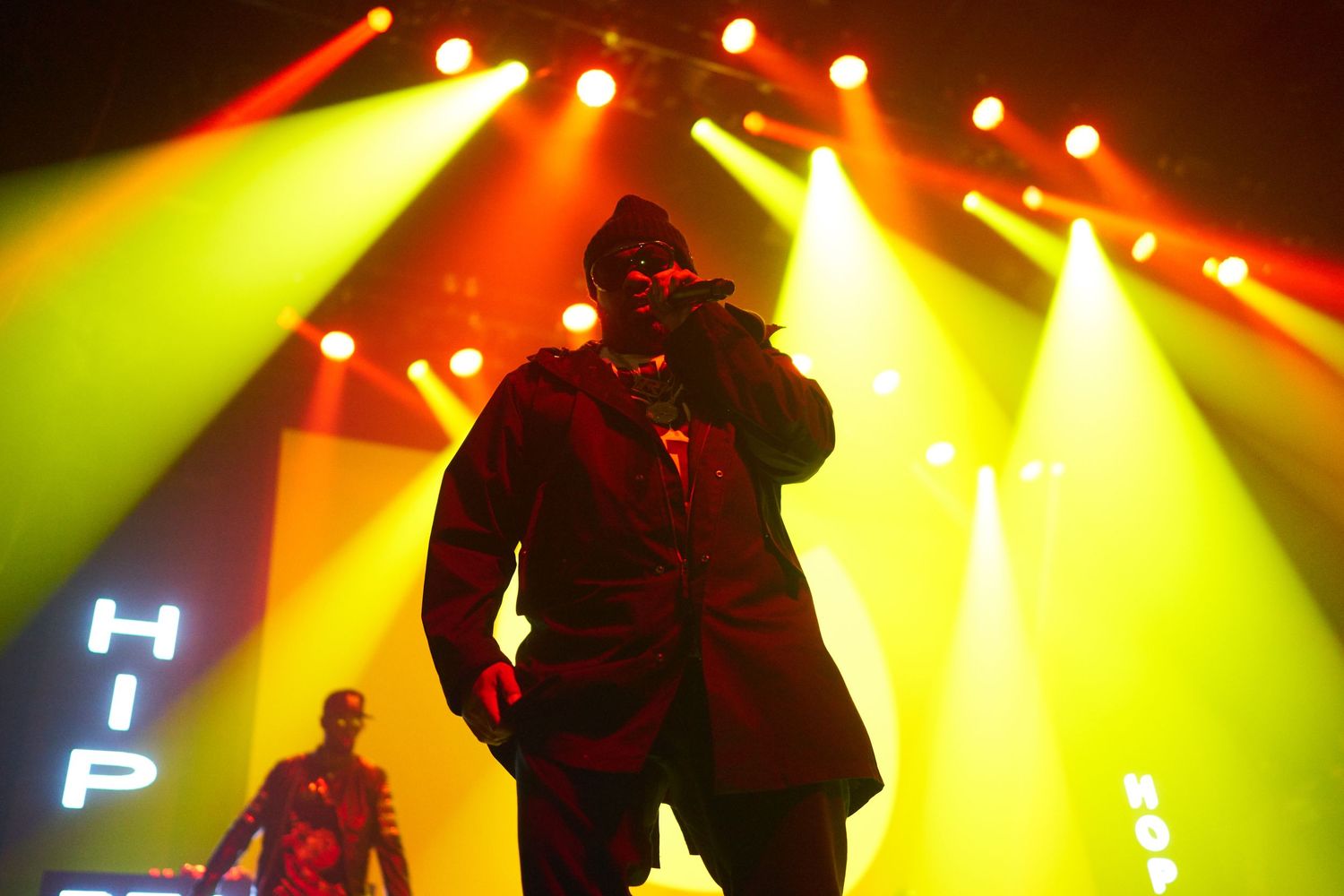 Silhouetted figures perform on stage with dramatic red and yellow concert lighting creating a vibrant atmosphere.