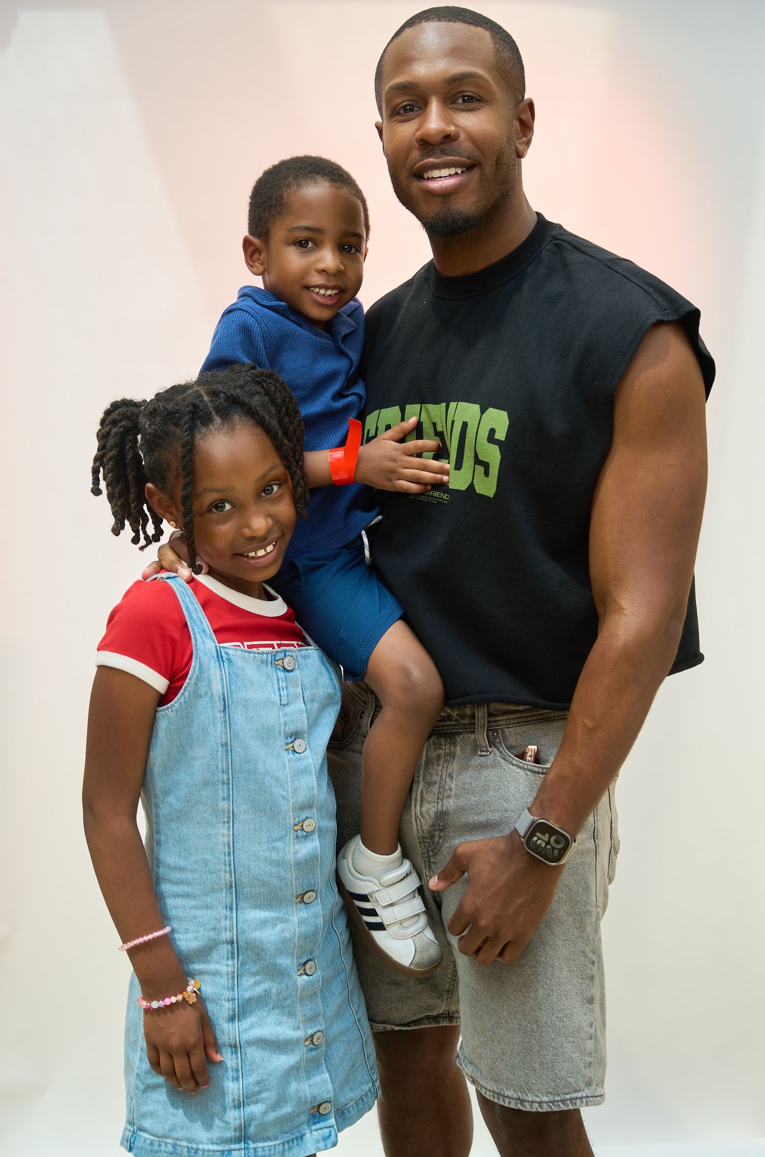 A man in a black sleeveless shirt holds a young boy while a girl in a denim dress smiles beside them.