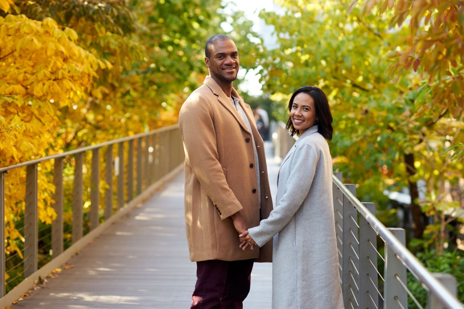 A couple holds hands while standing on a bridge surrounded by fall foliage with golden yellow leaves.