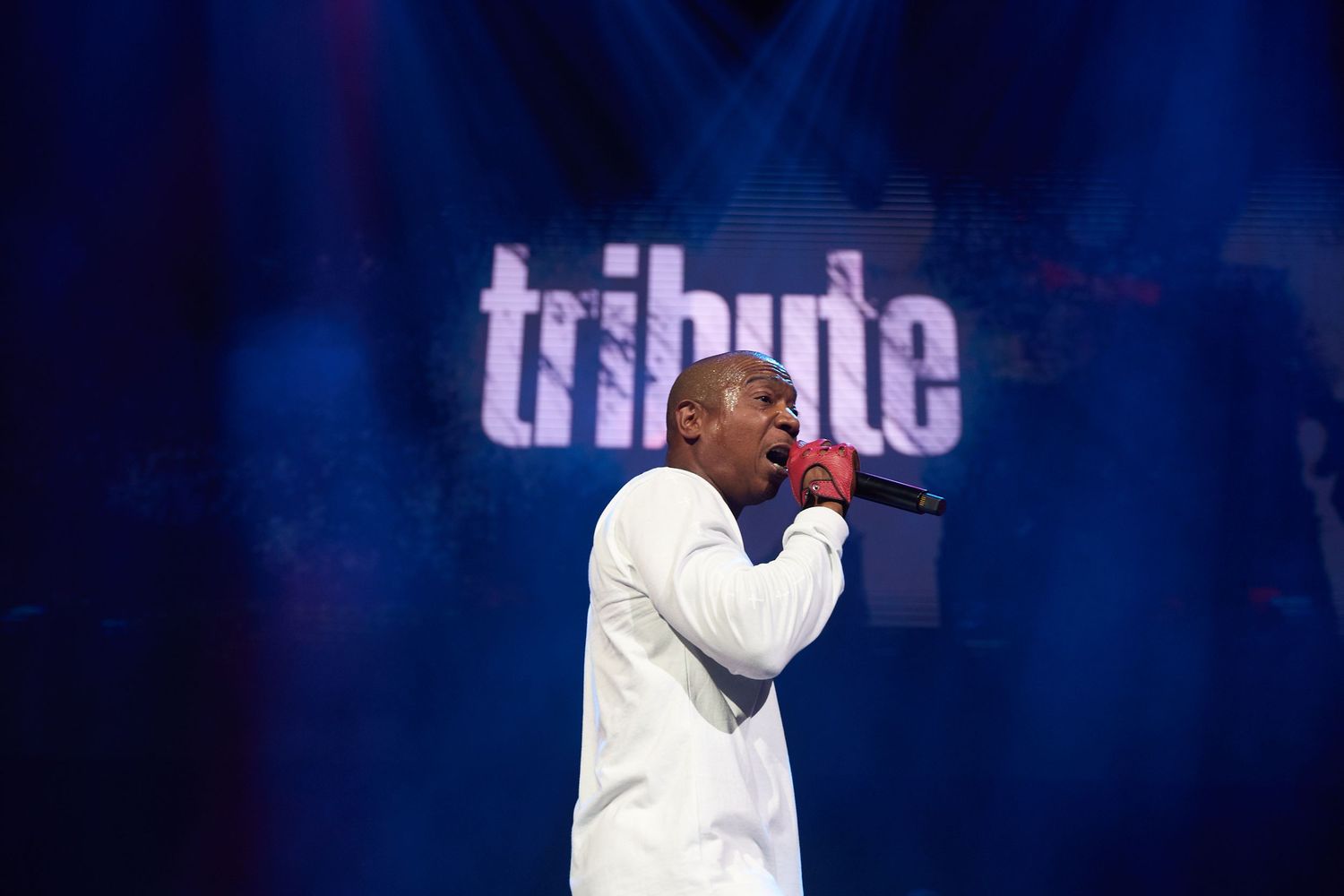 Performer in white outfit singing on stage with blue lighting and 'tribute' text display in background.