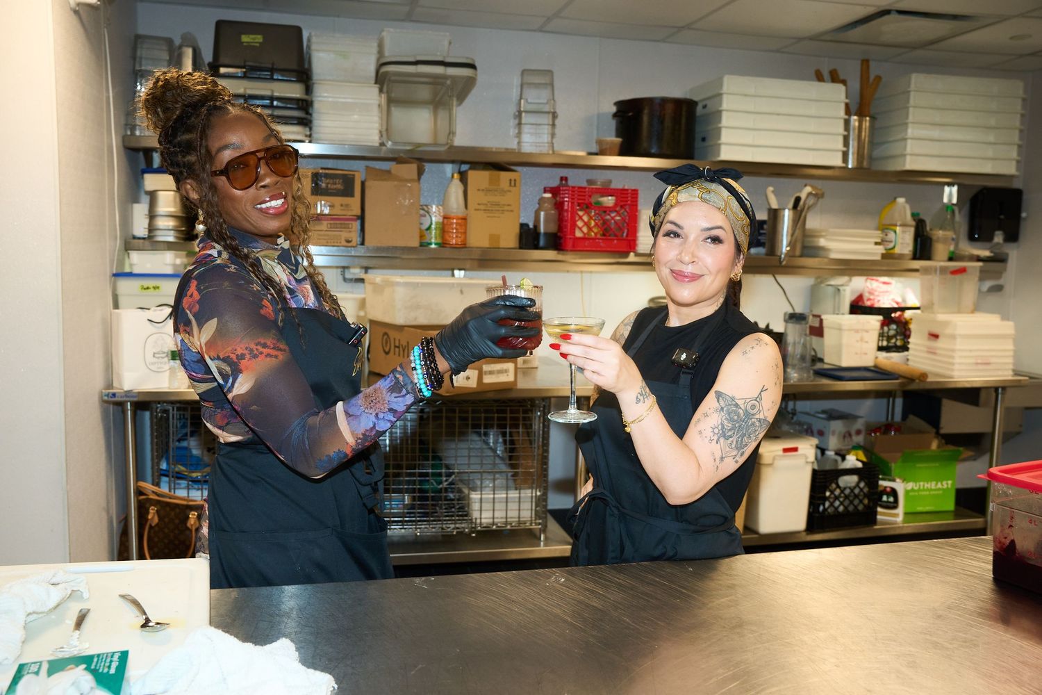 Two smiling women in black aprons raise cocktail glasses in a bustling commercial kitchen with stocked shelves and stainless steel counters.