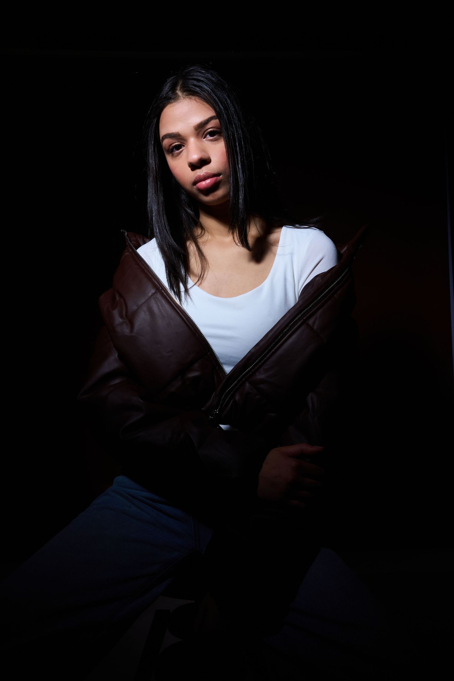 Dramatic portrait of a person wearing a white top and brown jacket against a dark background.