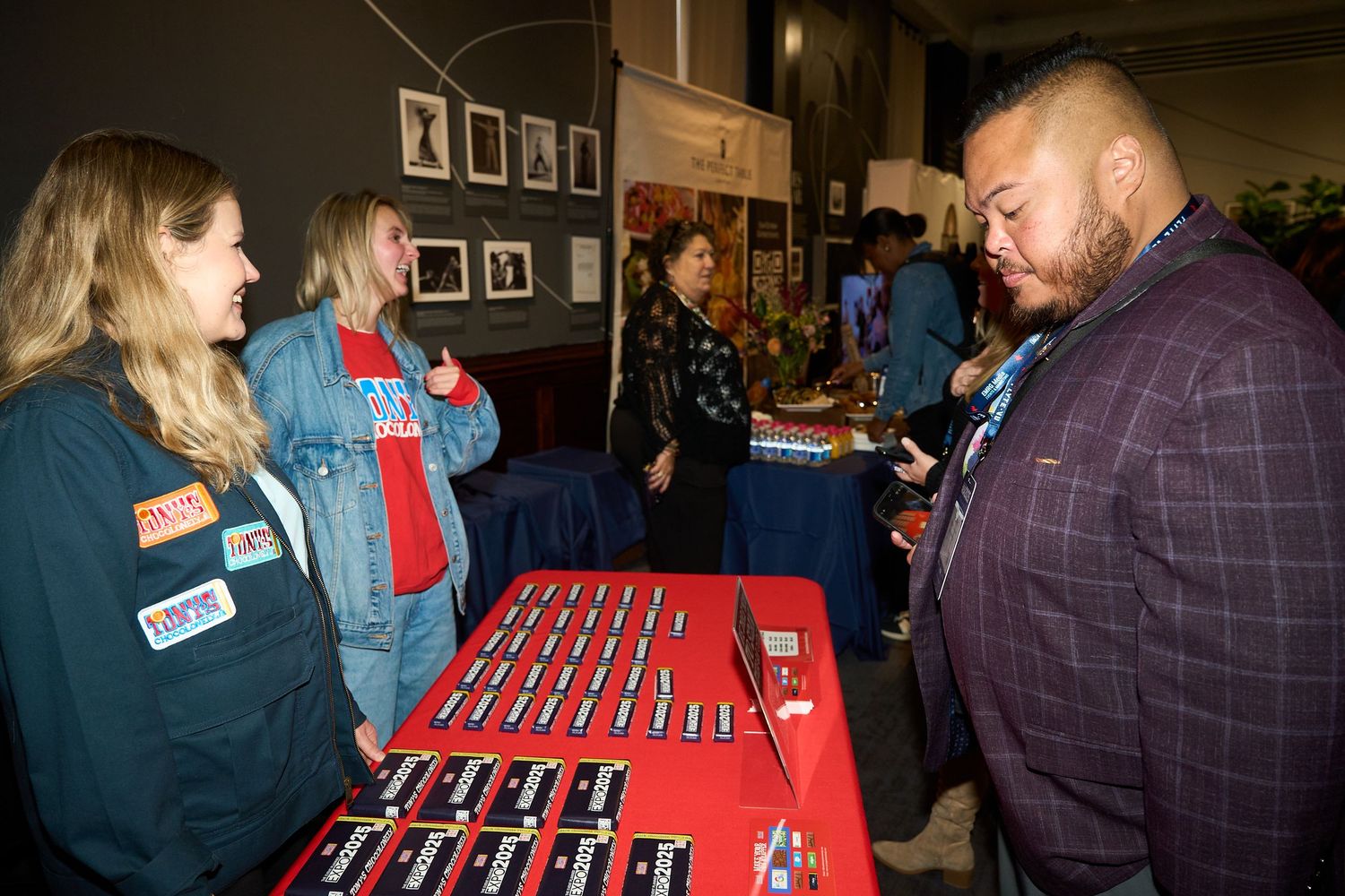 People gathered around tables at a social event signing papers or documents on red tablecloths.