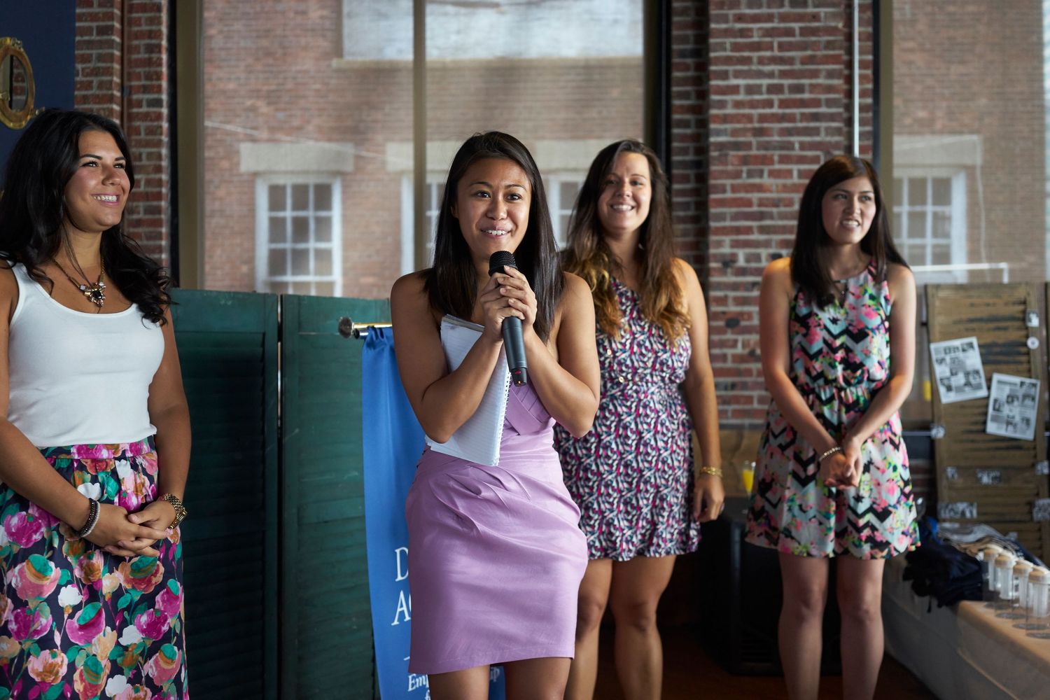 A group of people stand casually against a brick wall on what appears to be a school or university campus.