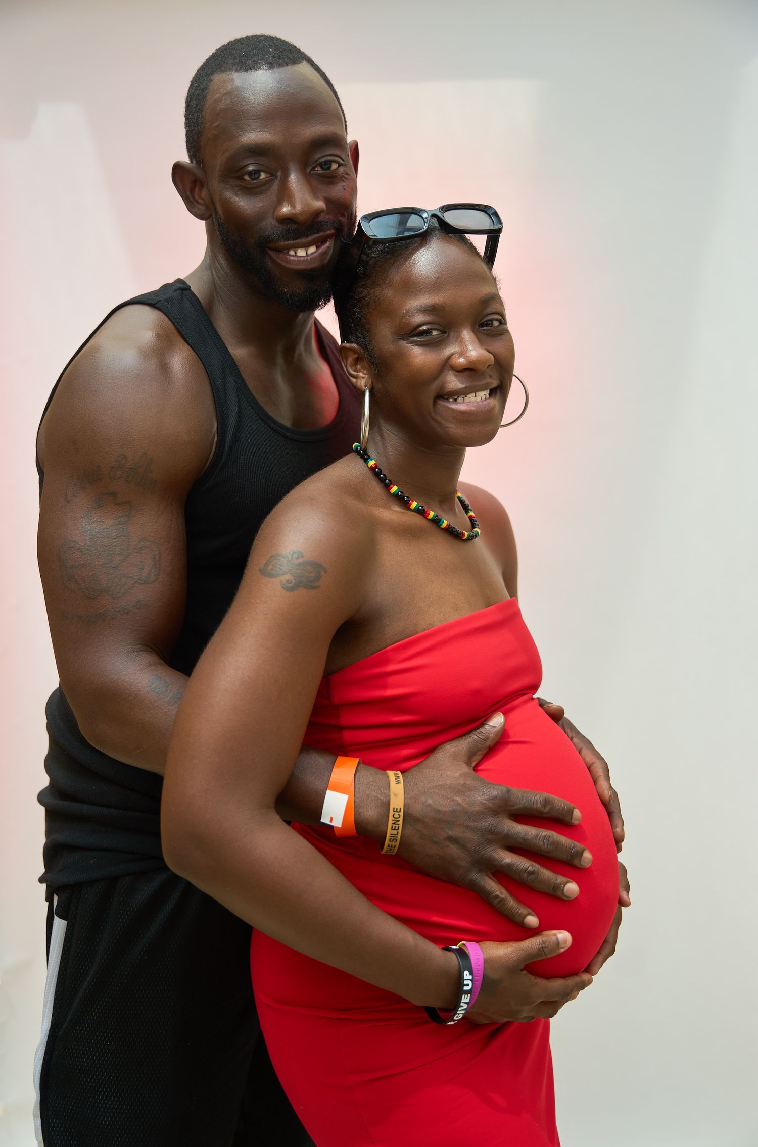 A smiling couple posing together, man in black tank top embracing pregnant woman in red strapless dress.