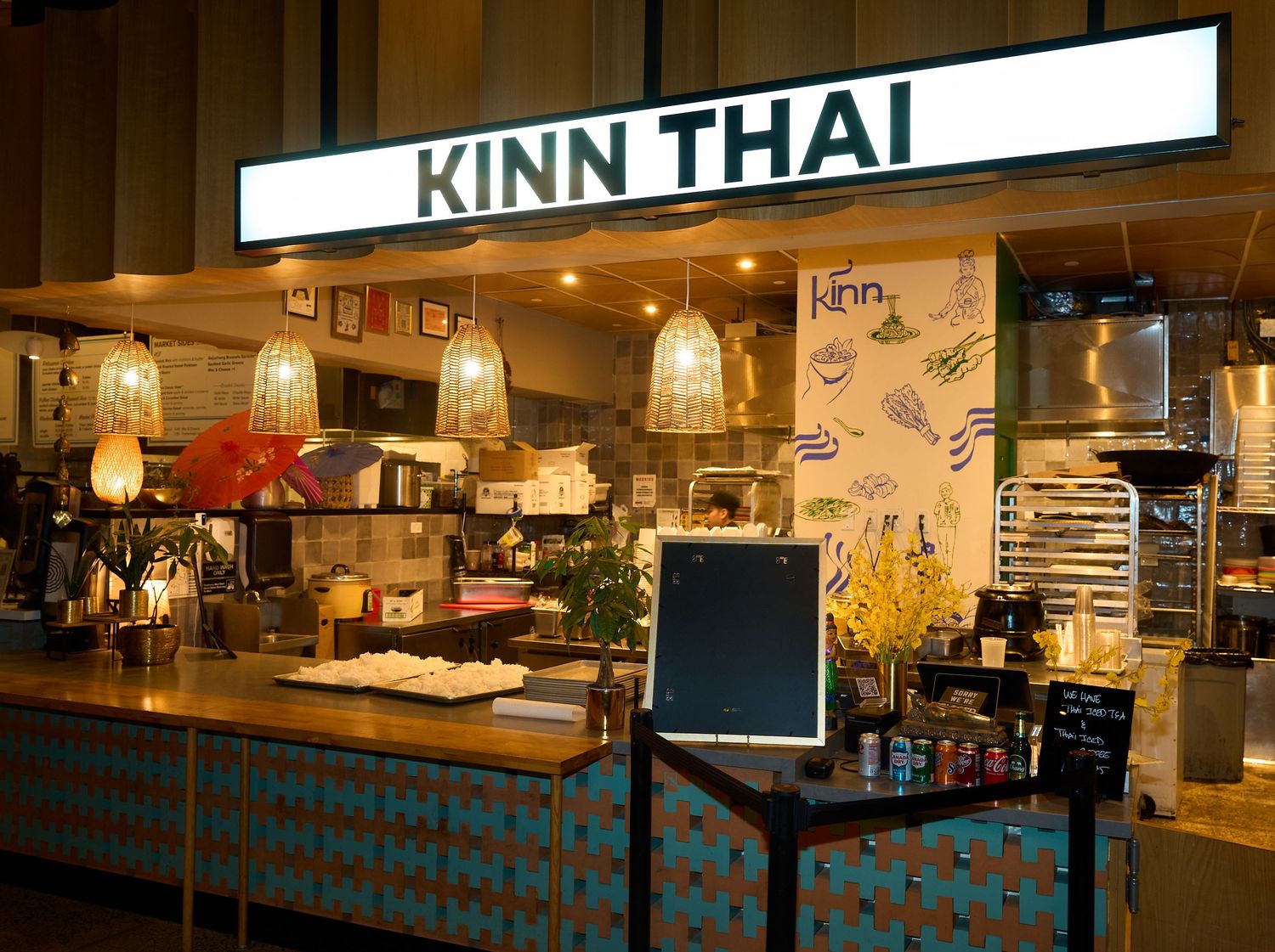 Kinn Thai restaurant counter with illuminated sign, pendant lamps, teal tiled front, and menu board in a warm, modern food hall setting.