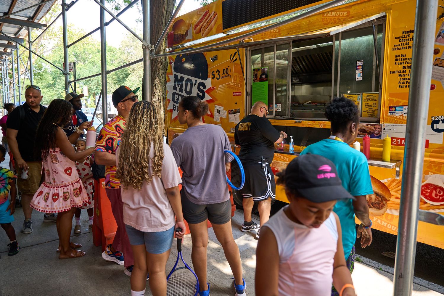 Customers line up at a yellow food truck parked under a covered area on a sunny day.
