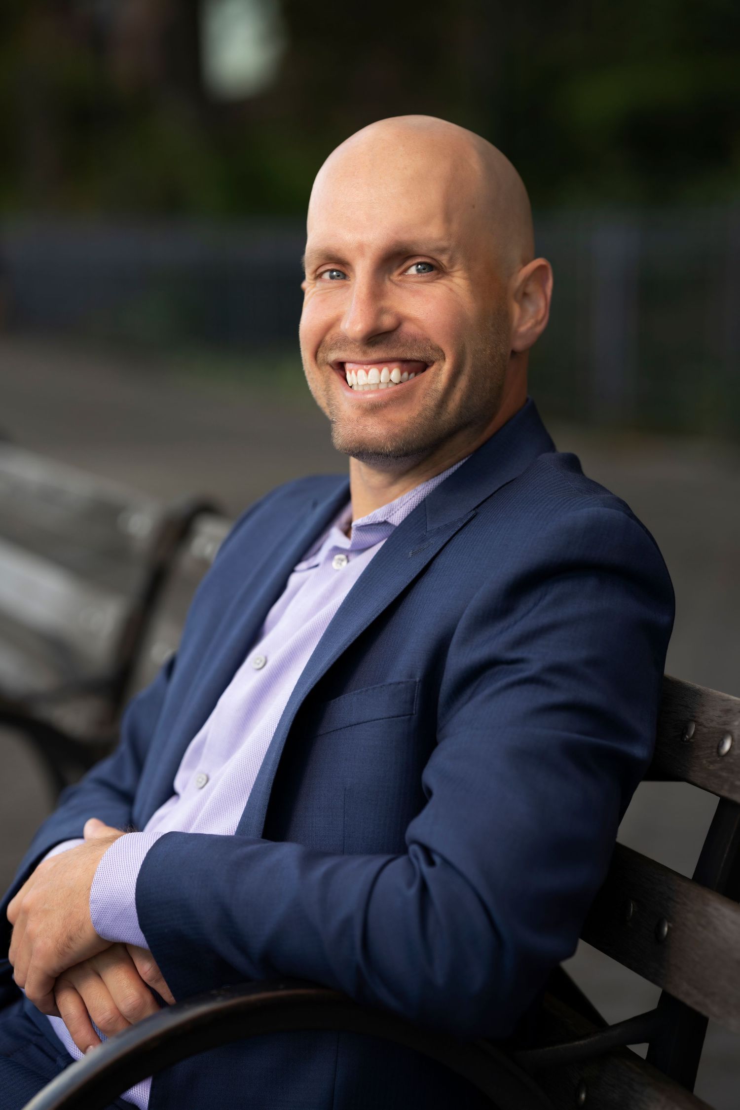 Professional headshot of a bald person in a navy suit jacket and light purple shirt smiling while seated outdoors.