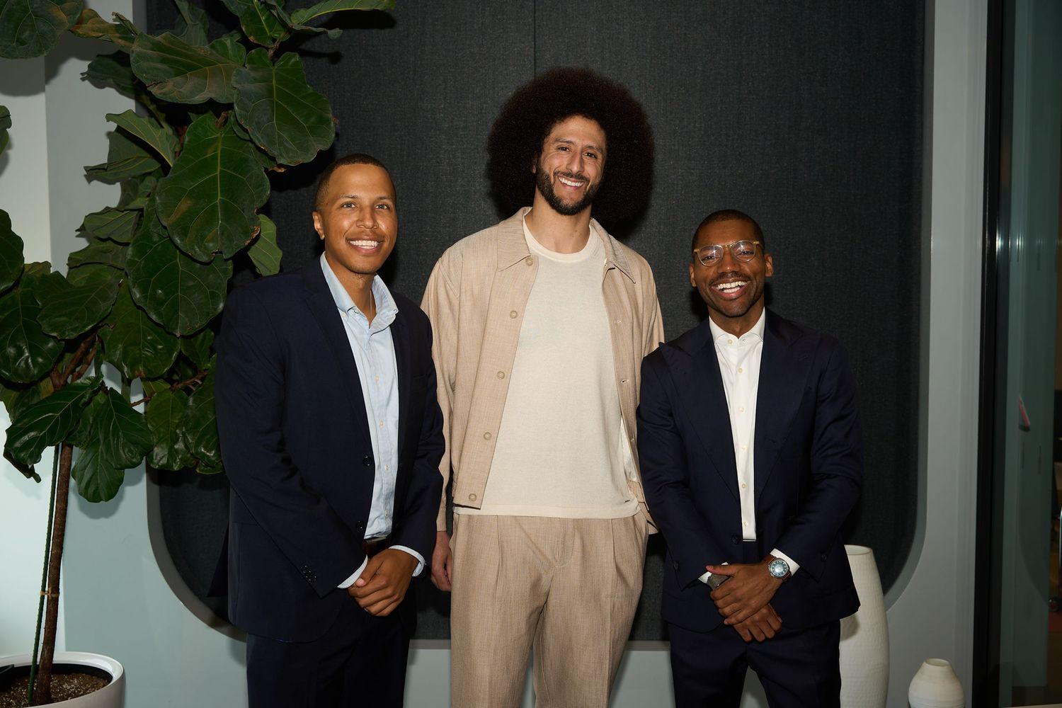 Three men in business casual attire stand together smiling against a dark wall with indoor plants.