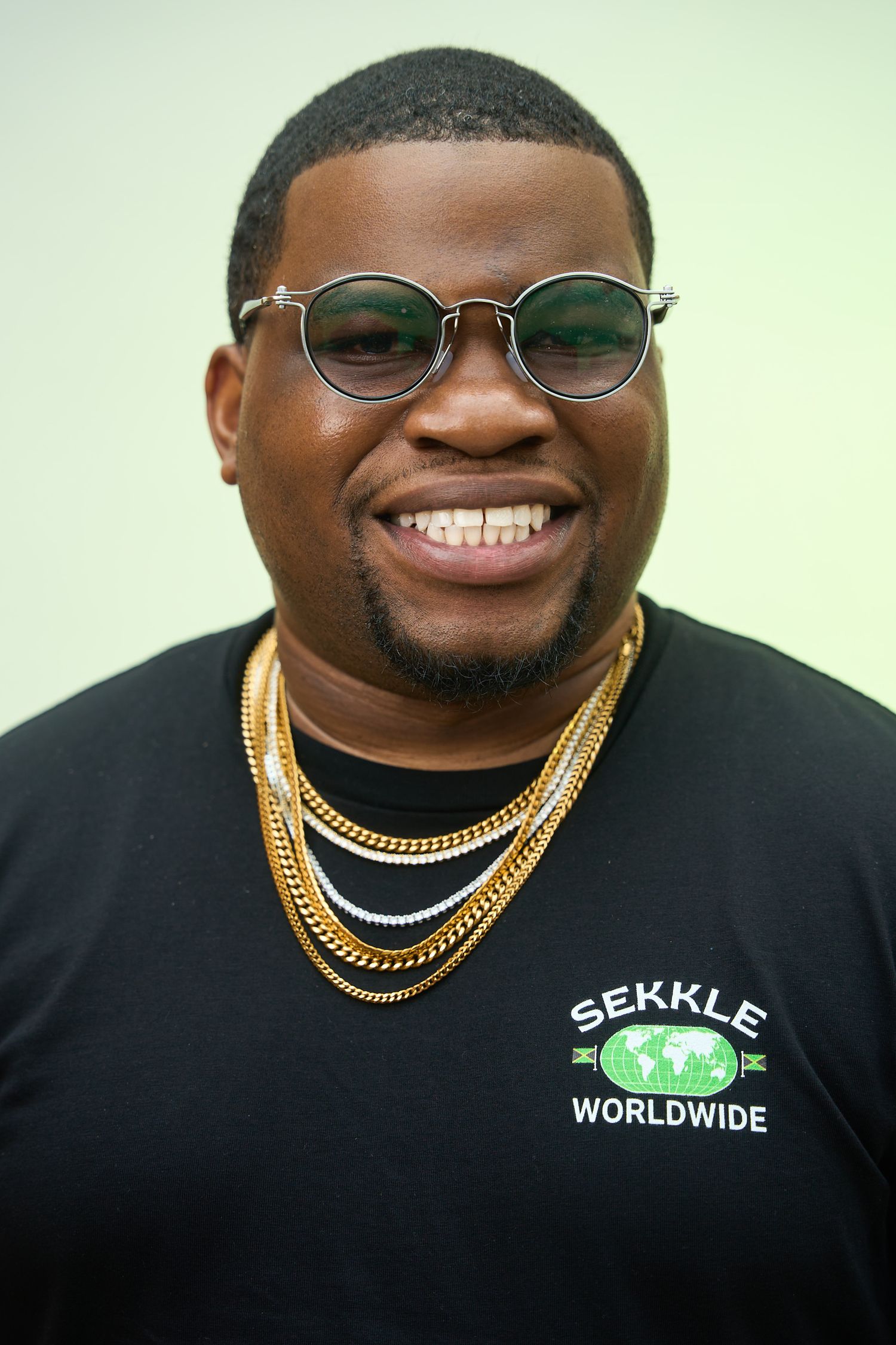 Smiling man wearing round glasses, gold chains, and a black Sekkle Worldwide t-shirt against a light green background.