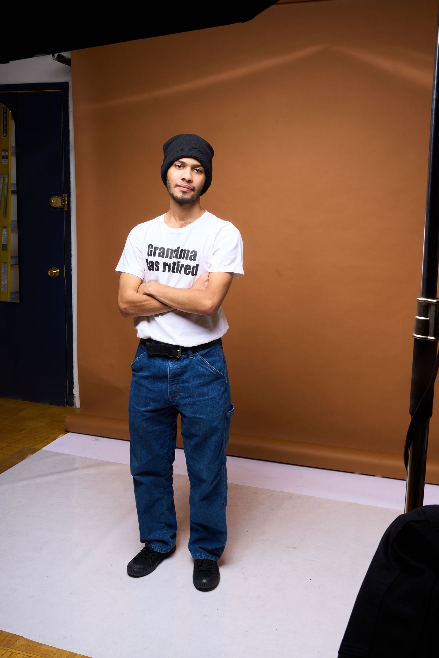 A person wearing a white t-shirt and blue jeans sits on a white floor against a brown backdrop in a studio setting.