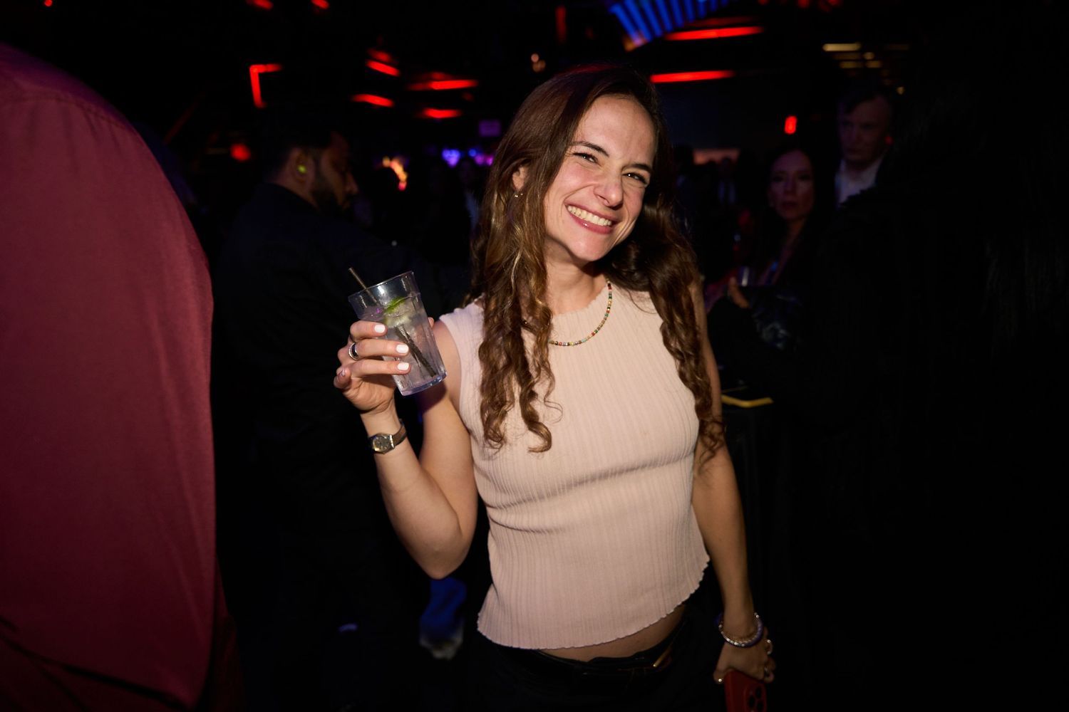 A person in a beige top smiling while holding a drink at a dimly lit party venue.