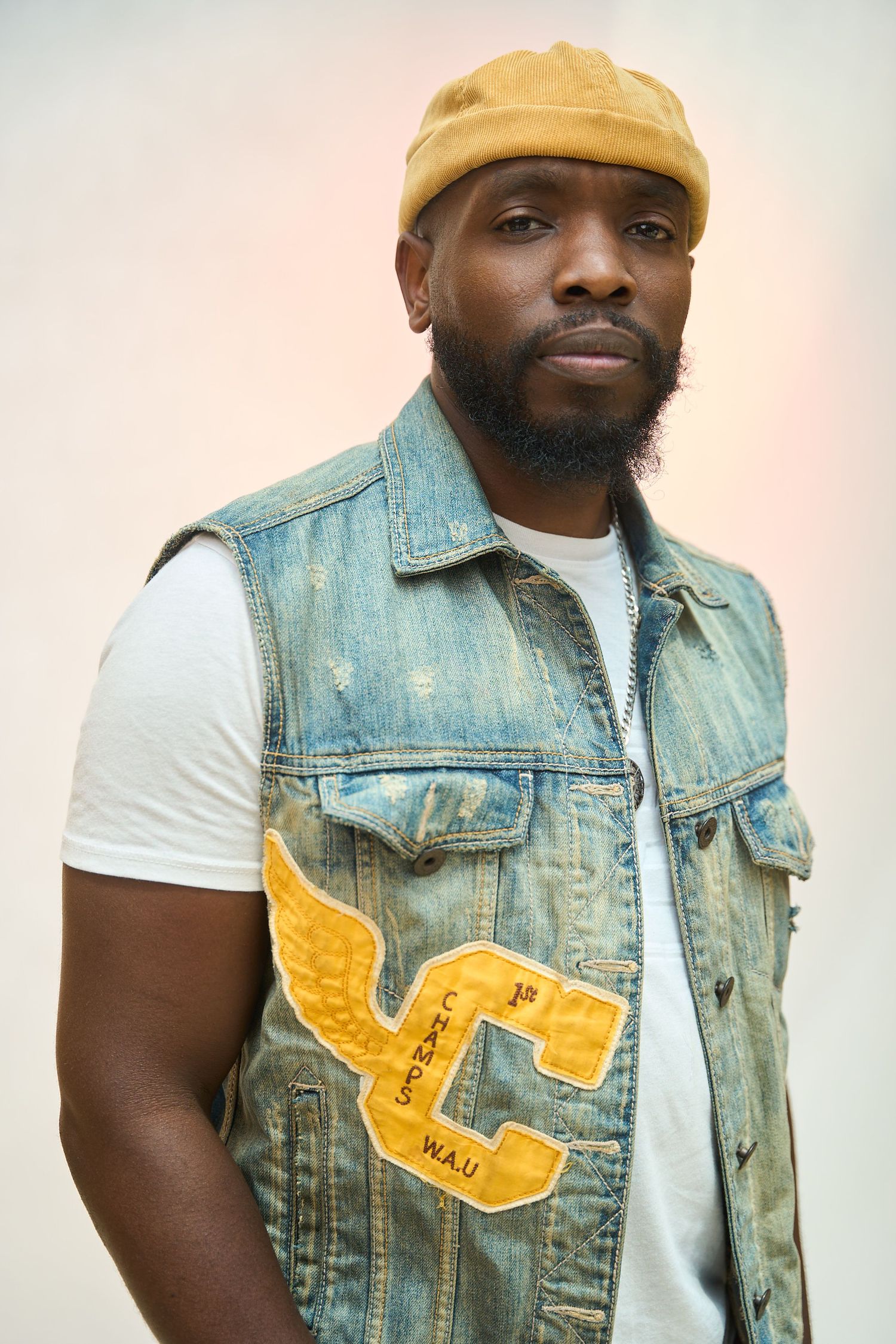 A bearded Black man wearing a yellow cap and denim vest with gold patches poses confidently outdoors.