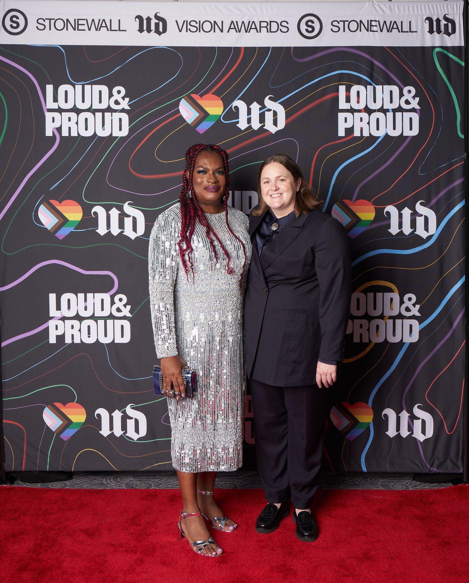 Two attendees pose on red carpet at Stonewall ITN Vision Awards event with Pride logos and decorative backdrop.