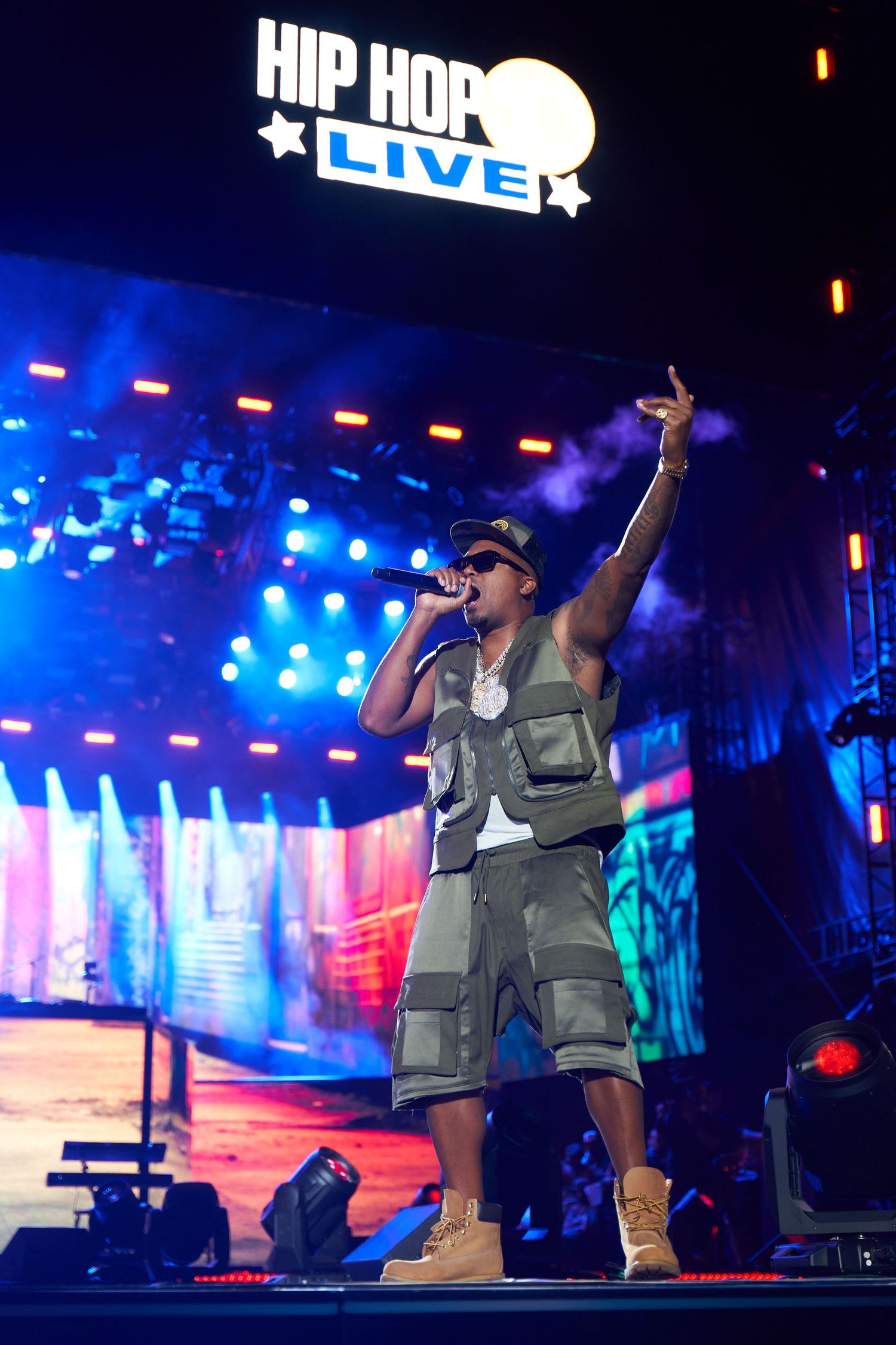 Hip hop performer wearing cargo vest energetically performs on illuminated stage with dramatic blue lighting effects.