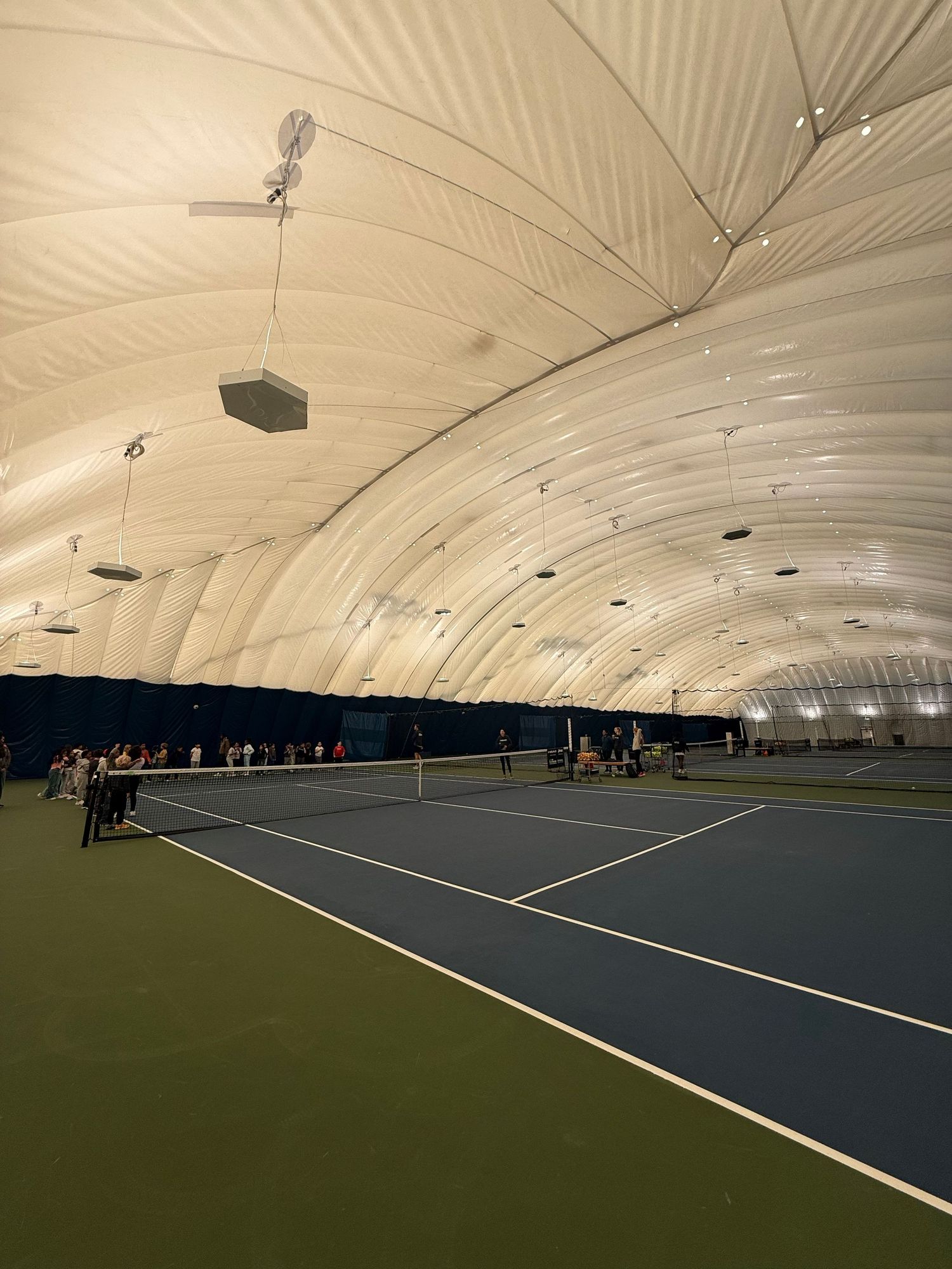 Indoor tennis facility with multiple courts under a large white dome structure showing various playing surfaces and lighting.