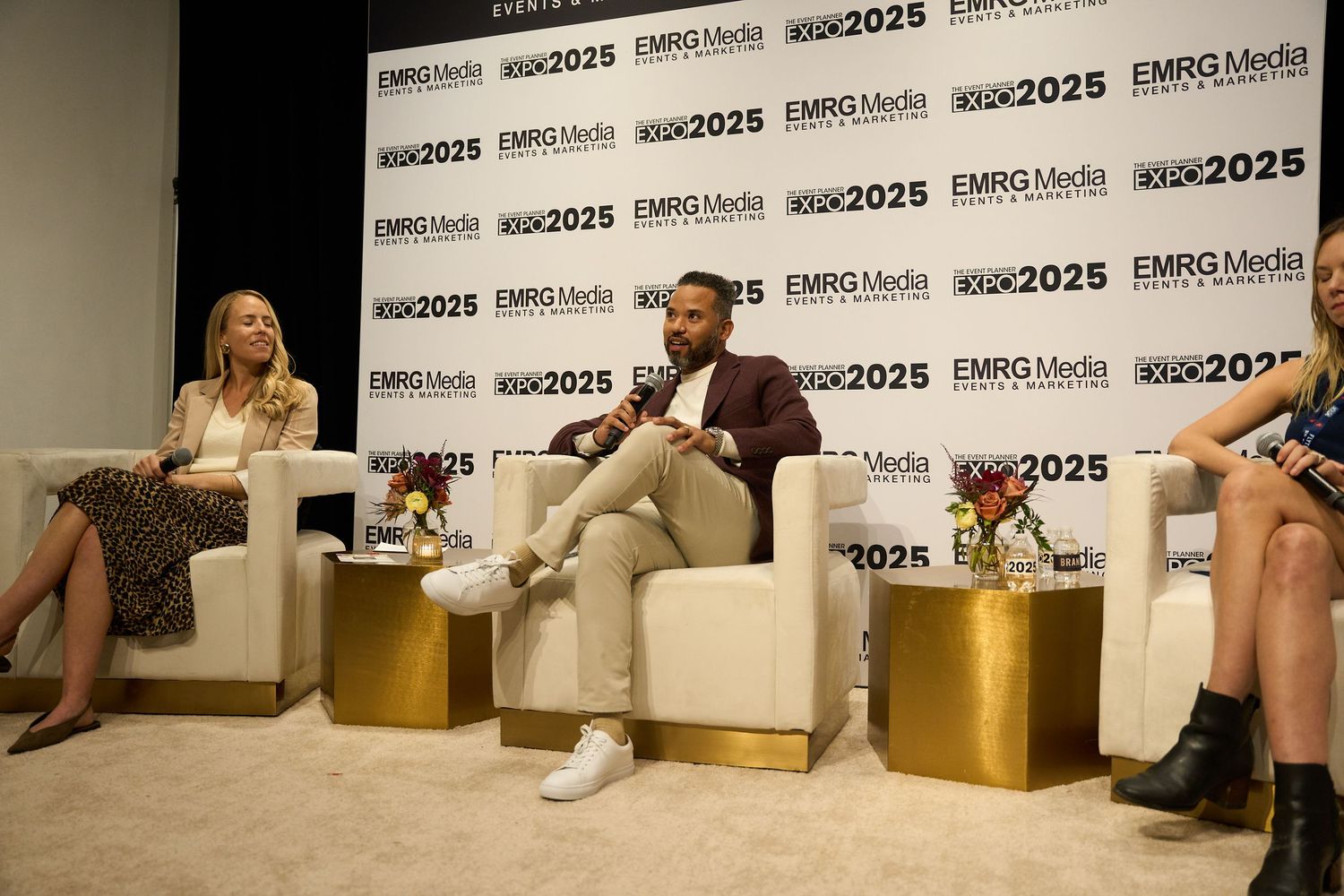 Panel discussion participants seated on white chairs and gold cubes on stage during a conference presentation.