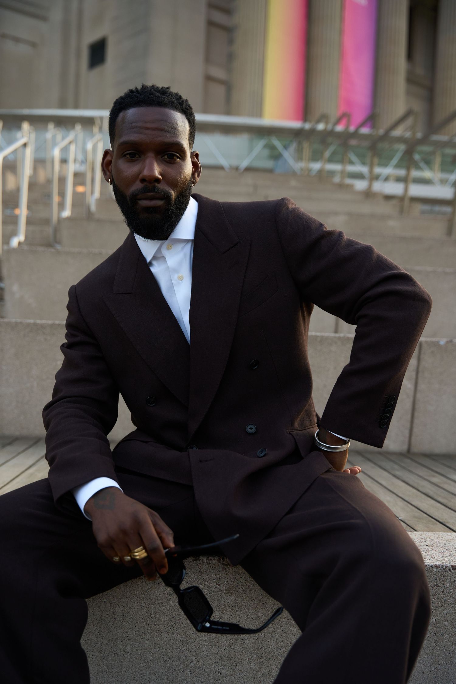 Well-dressed person in a dark suit and white shirt sits on concrete steps with soft pink lighting in background.