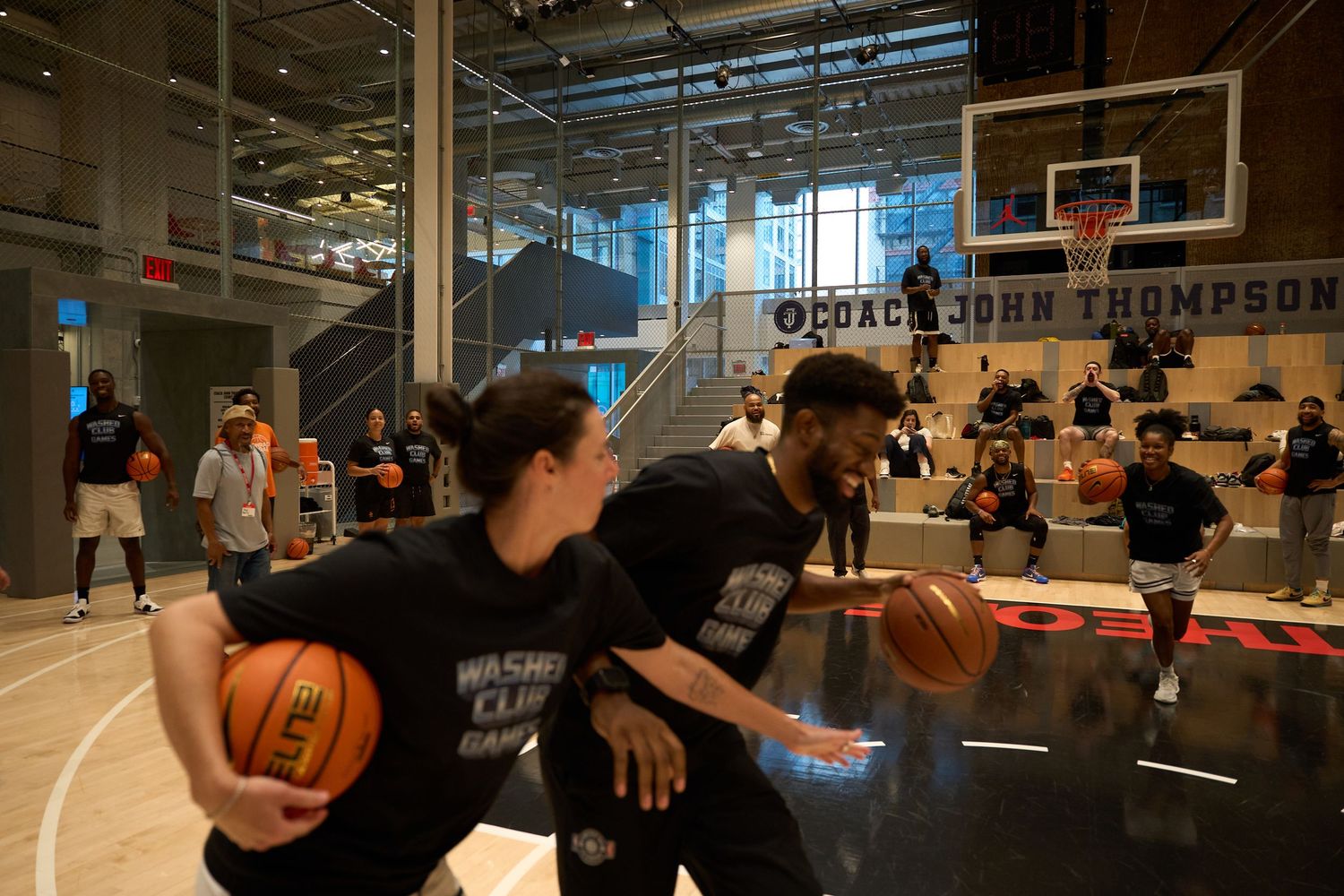 Basketball players practice dribbling drills on an indoor court with glass windows and basketball hoops visible.