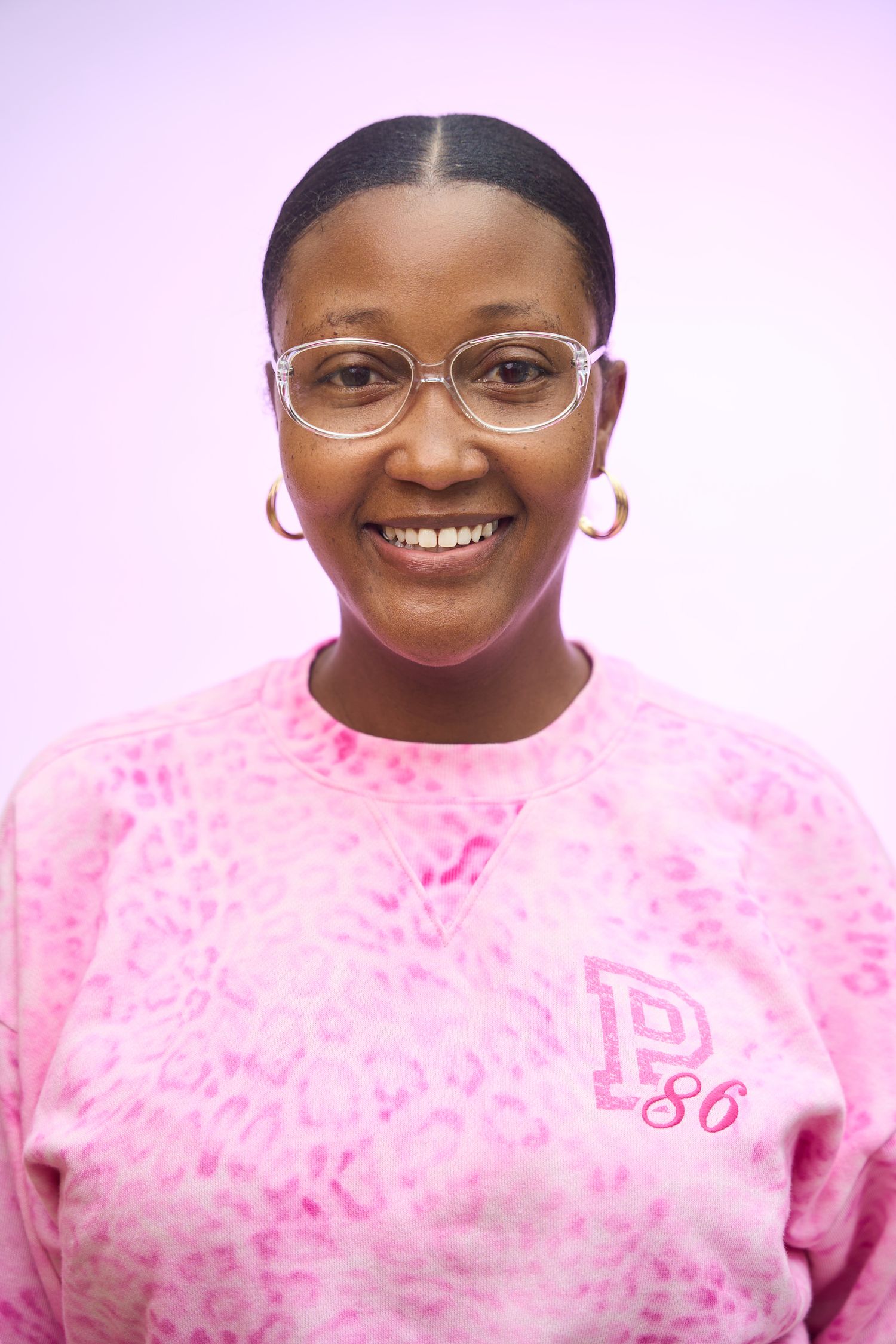 Smiling woman wearing pink tie-dye sweatshirt and clear glasses against a light pink background.