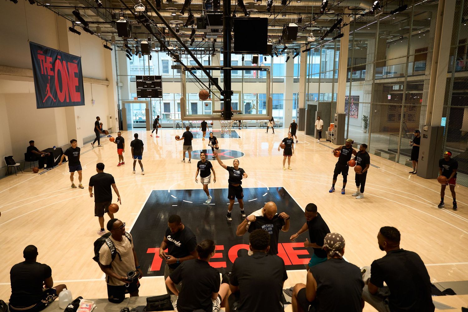 Players practicing basketball in a bright indoor court with spectators watching from bleachers.