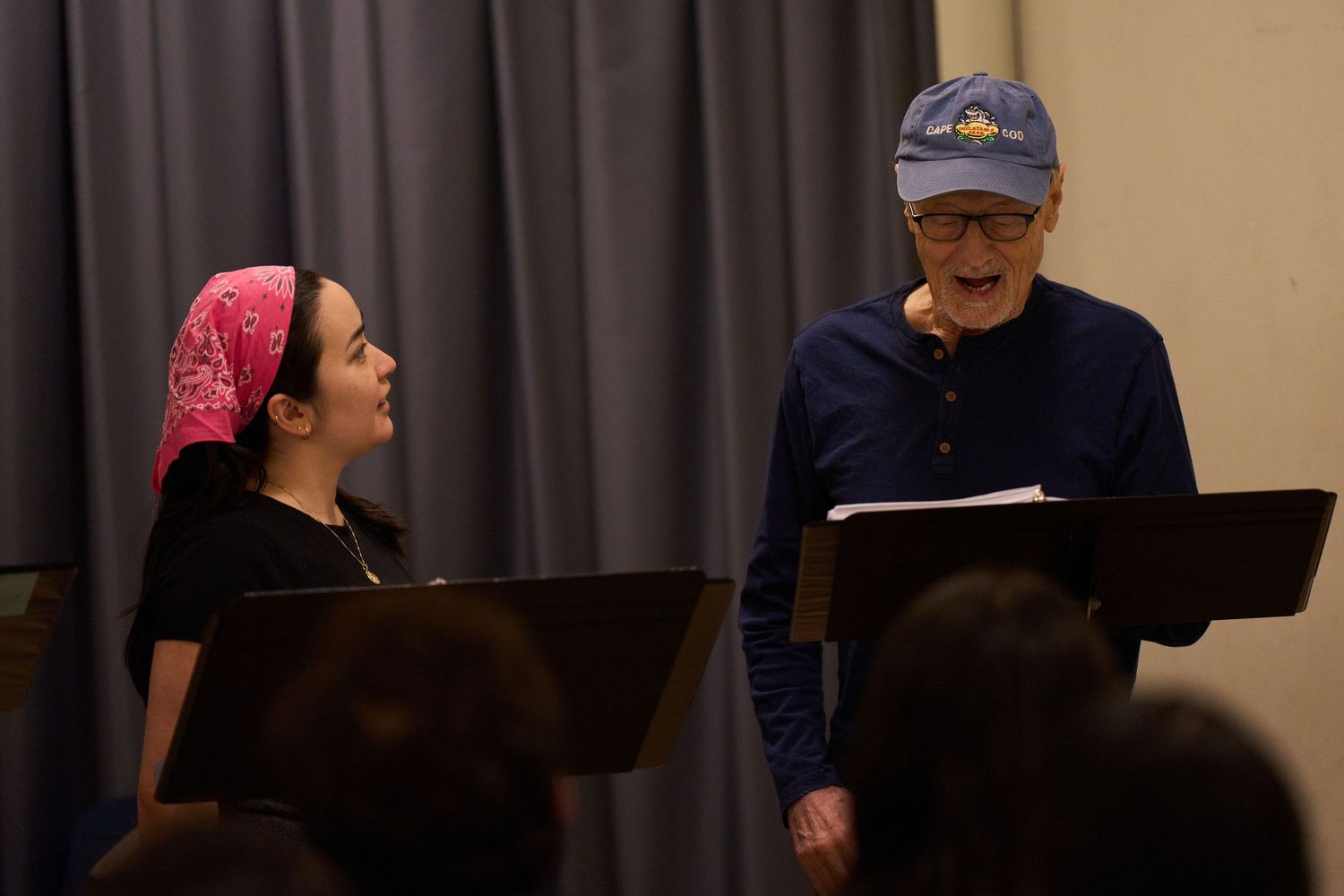 Two performers standing at music stands rehearse together on stage with one wearing a red bandana and black shirt.