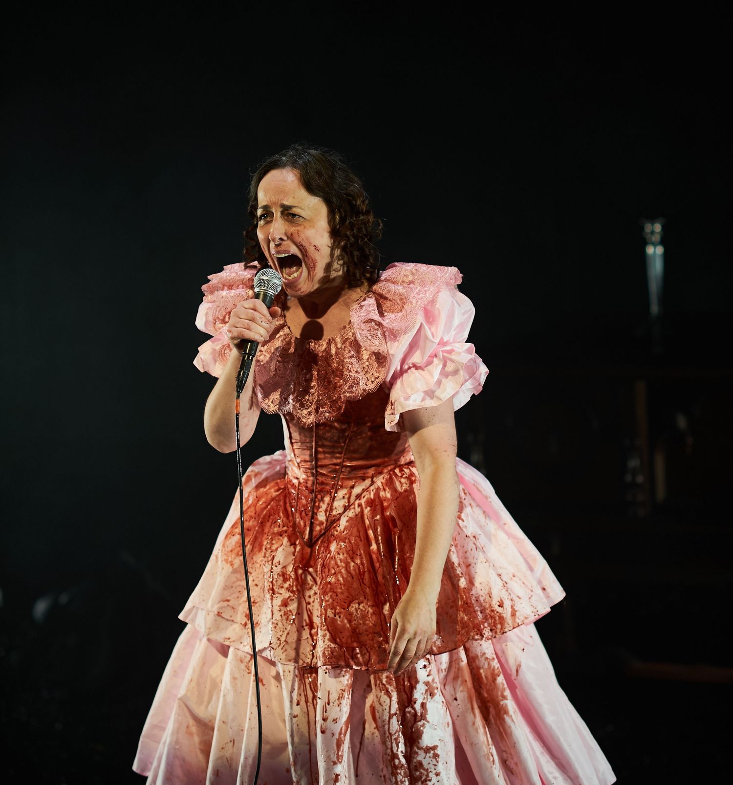 A performer in a blood-stained pink ruffled dress passionately sings on a dark stage during a dramatic theatrical number.